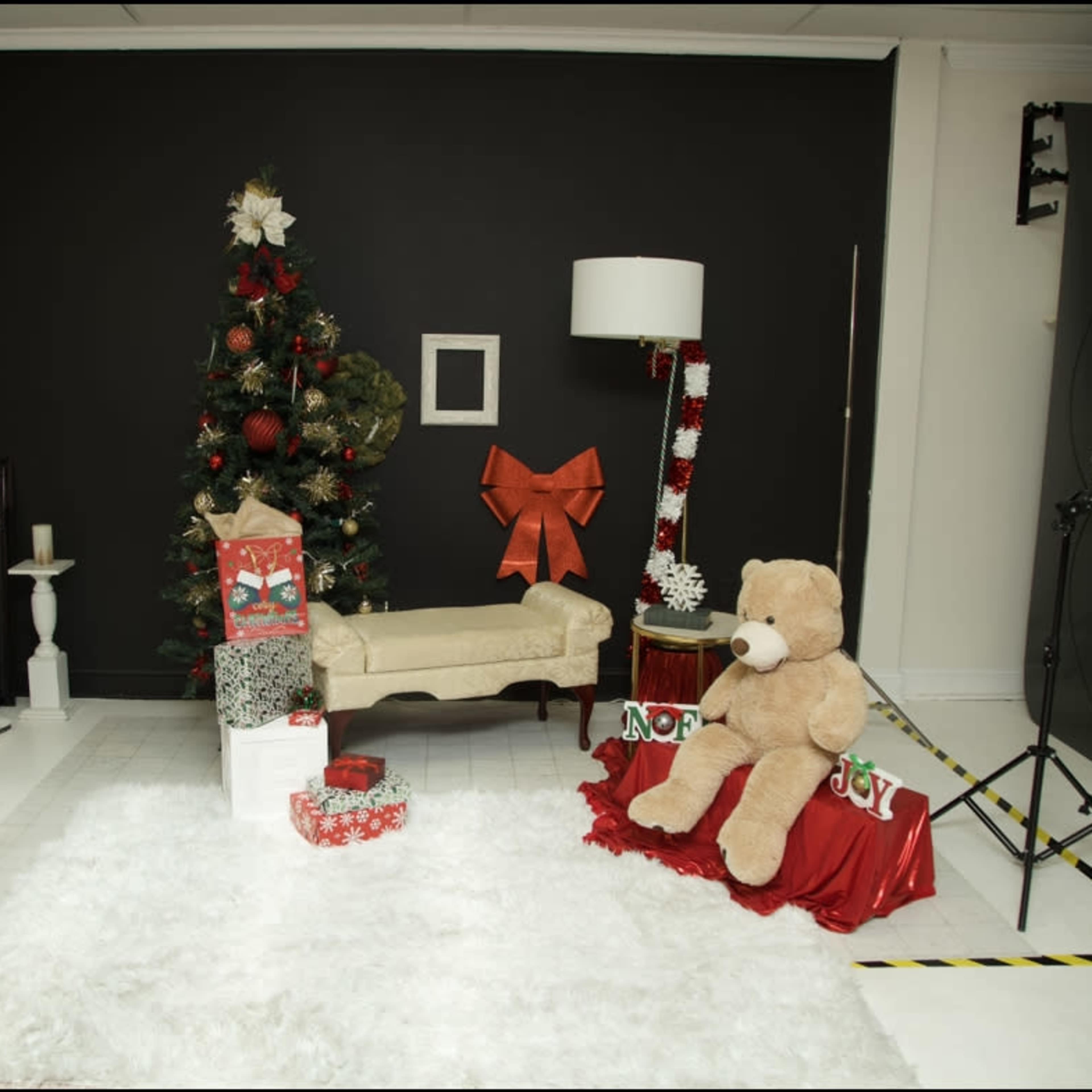 A decorated studio corner featuring a Christmas tree, a small couch, a large teddy bear, and wrapped gifts on a white rug.
