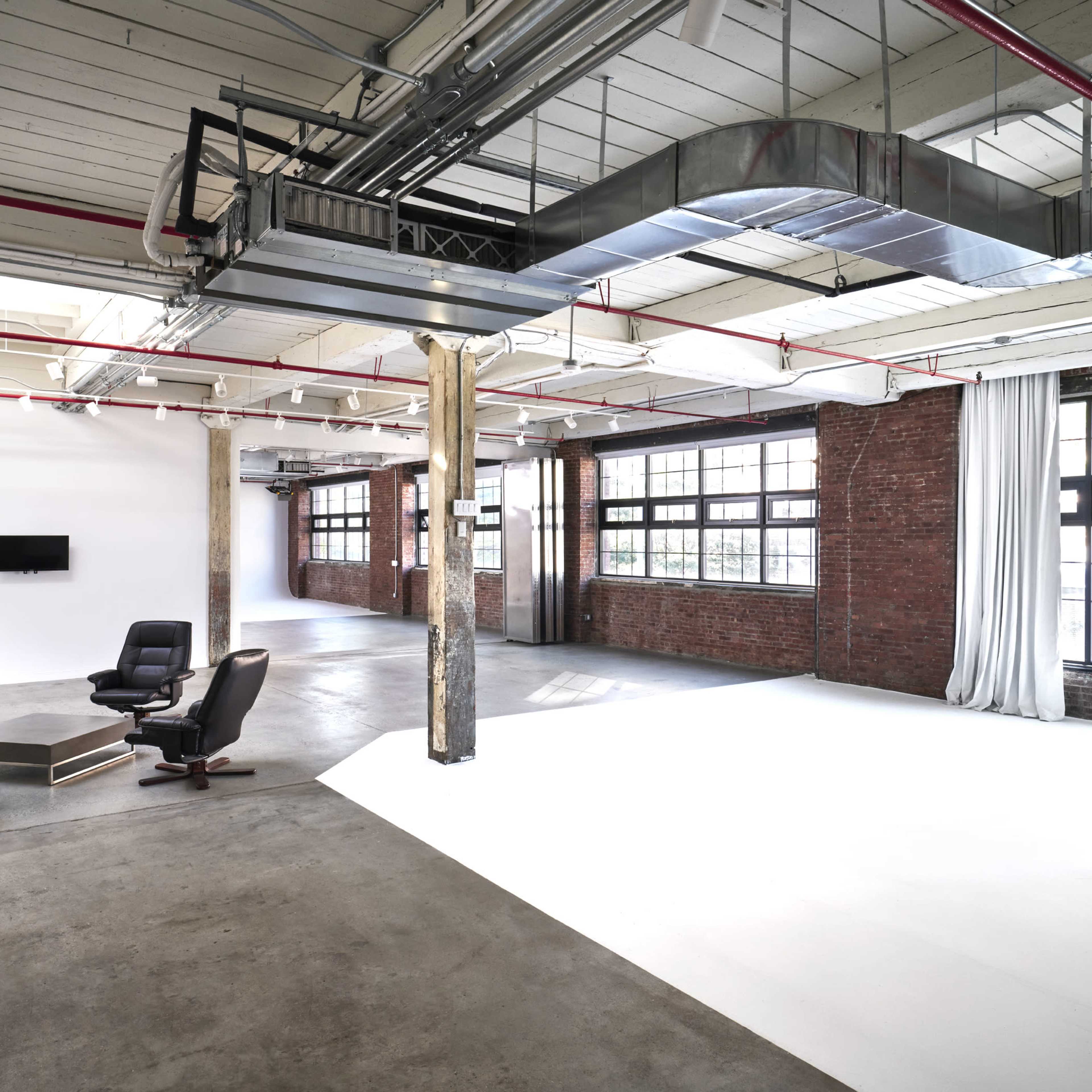An industrial-style interior space featuring four black chairs arranged in a circle, large windows allowing natural light, and exposed brick and concrete elements.