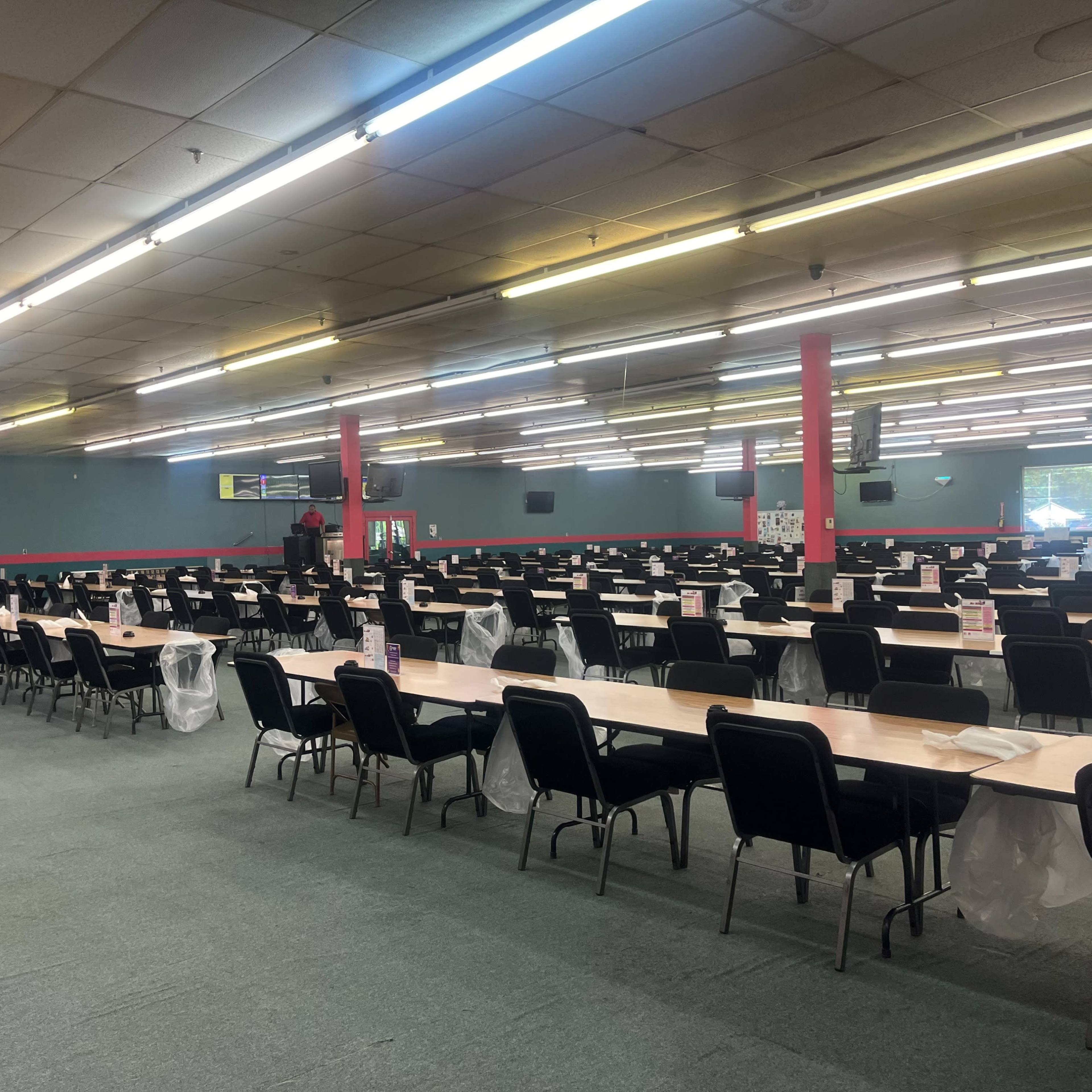 A large hall is filled with rows of long tables and chairs, illuminated by overhead lights.