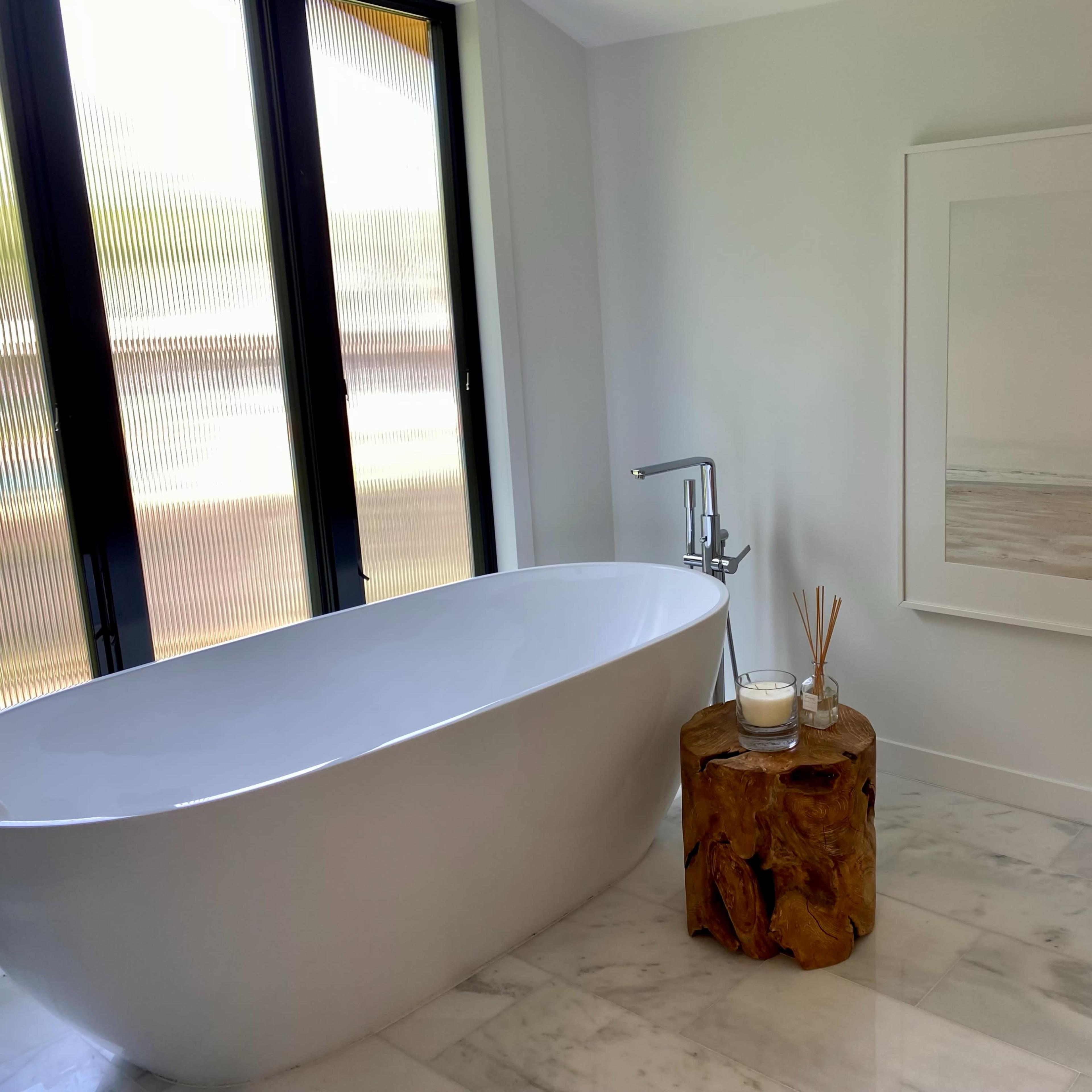 A modern bathroom featuring a freestanding white bathtub, a wooden side table, and a large framed artwork on the wall.