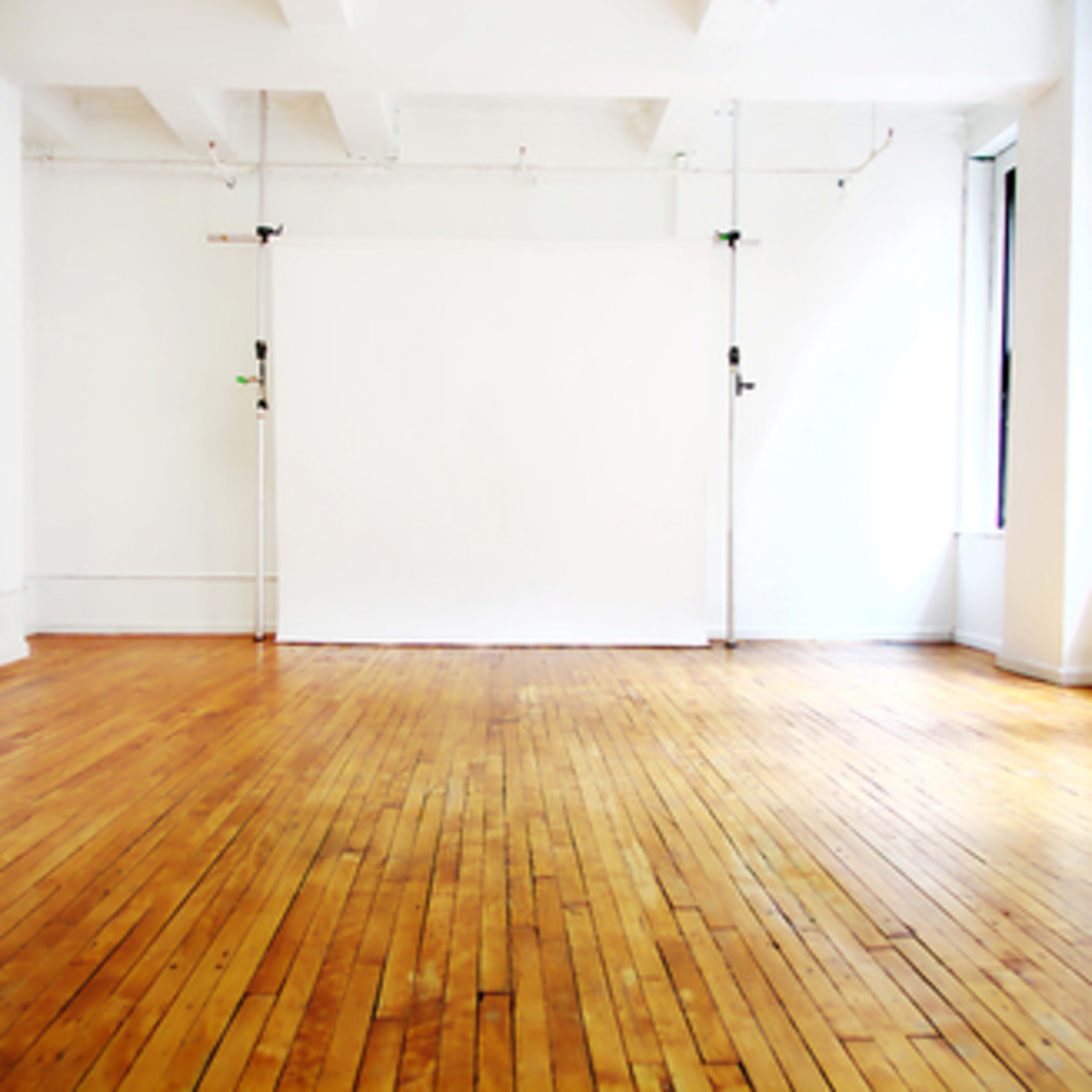 A bright, empty studio space features polished wooden floors and a blank white backdrop against the wall.