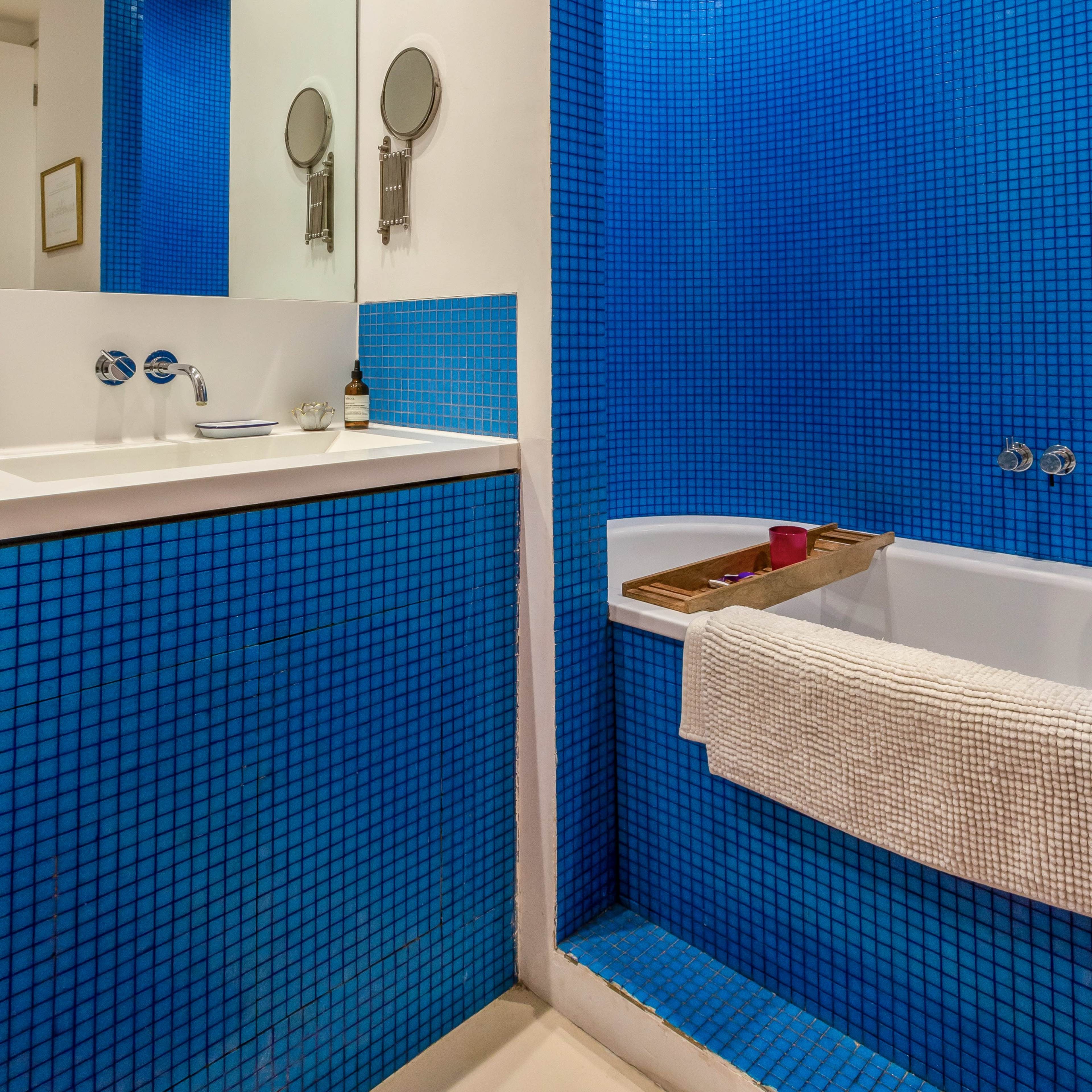 A modern bathroom with blue mosaic tile walls, a white countertop with sink fixtures, and a bathtub lined with a wooden tray.