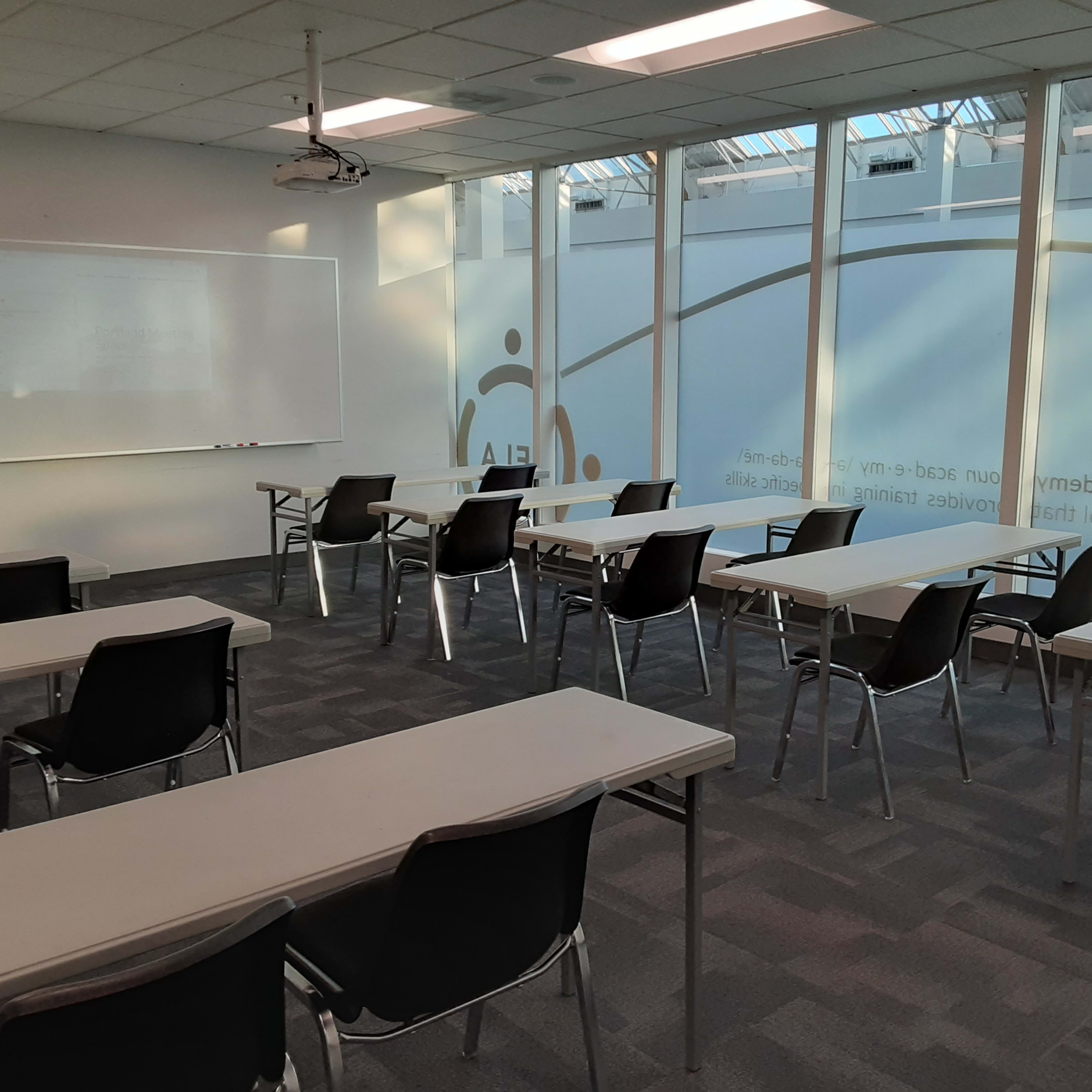 A classroom features several rows of tables and chairs, with large windows letting in natural light.