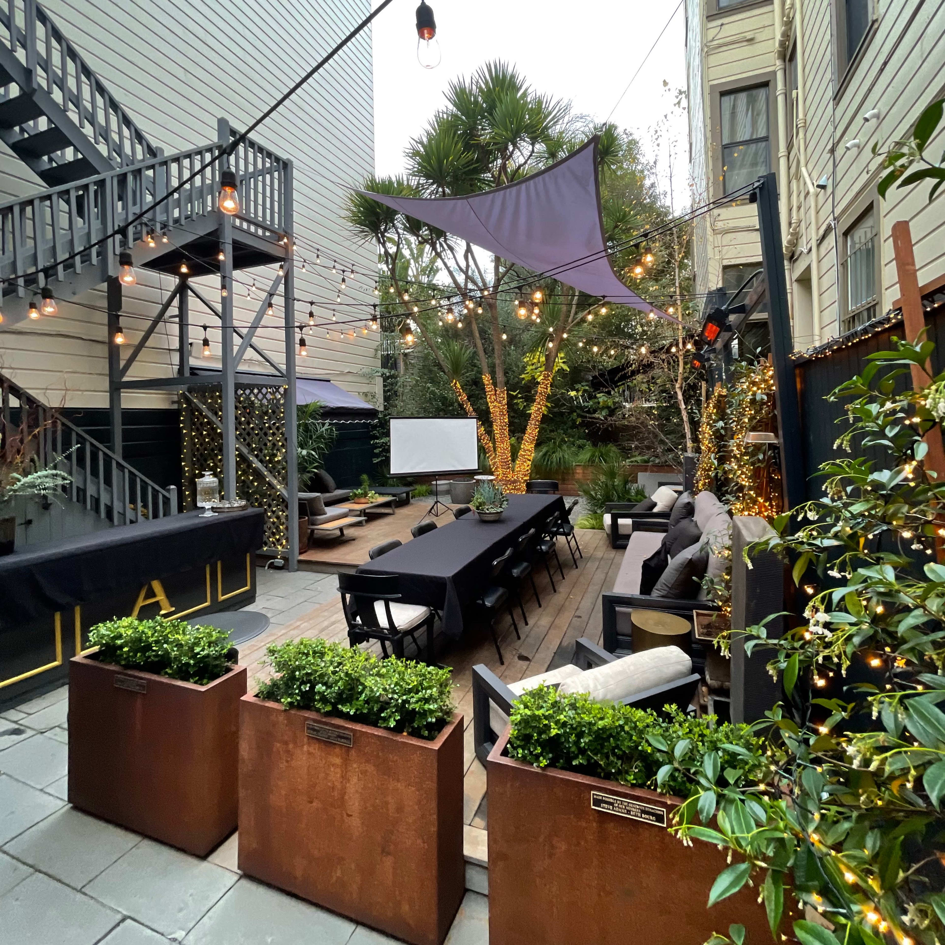 A courtyard with a long dining table surrounded by planters, string lights overhead, and a projector screen at one end.