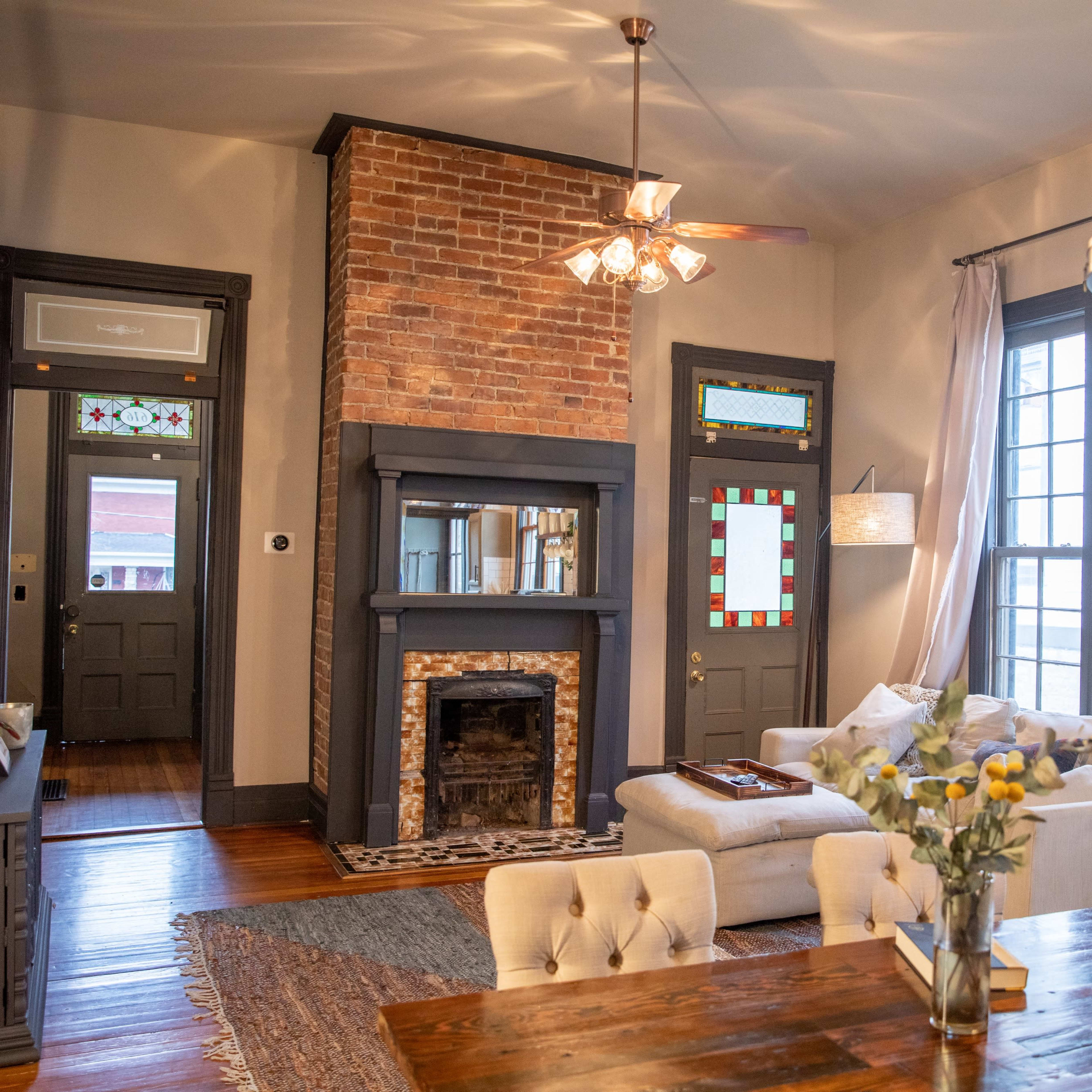 A cozy living room featuring a brick fireplace, light-colored furniture, and large windows with decorative stained glass.