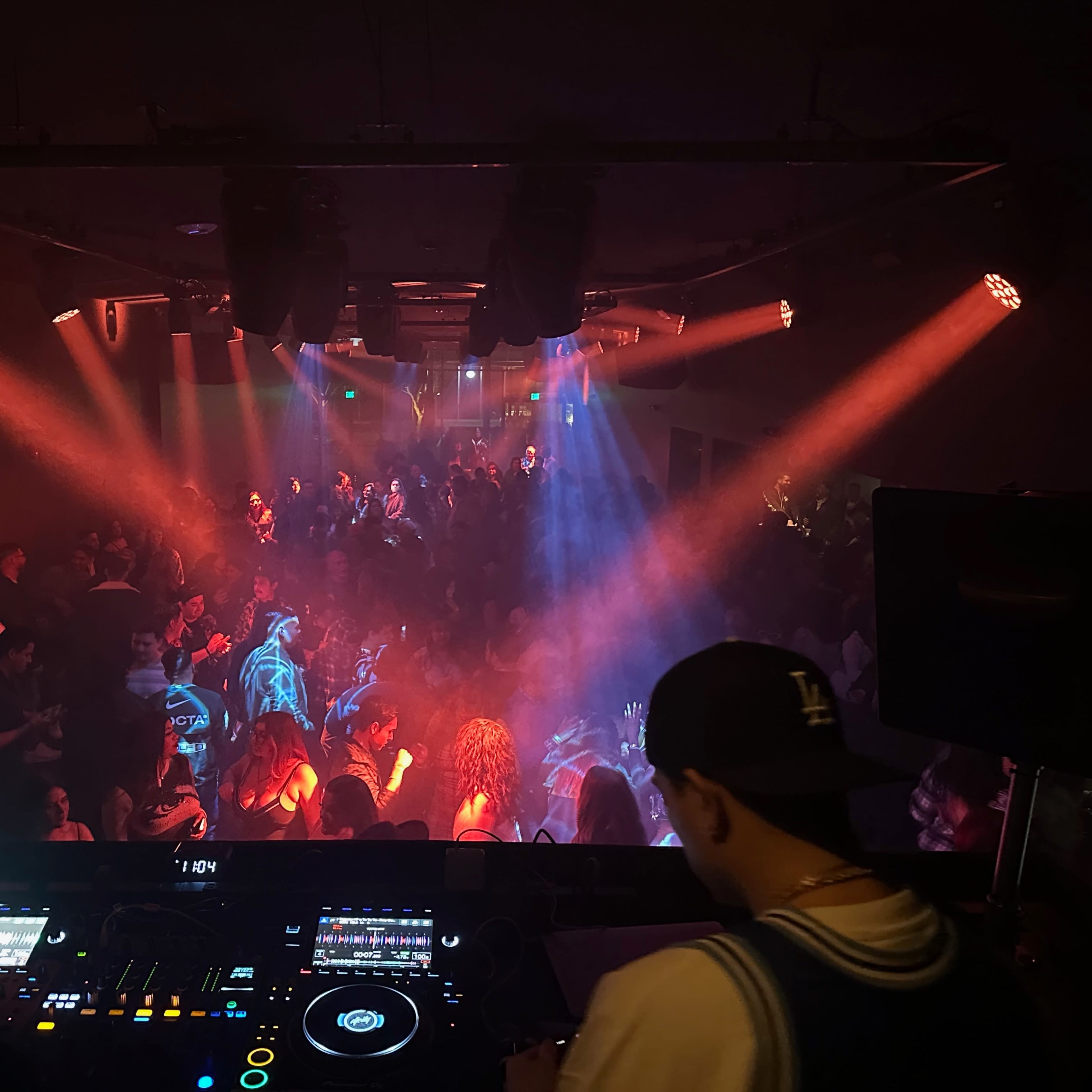A DJ performs in a club while a crowd dances under colorful lights and fog.