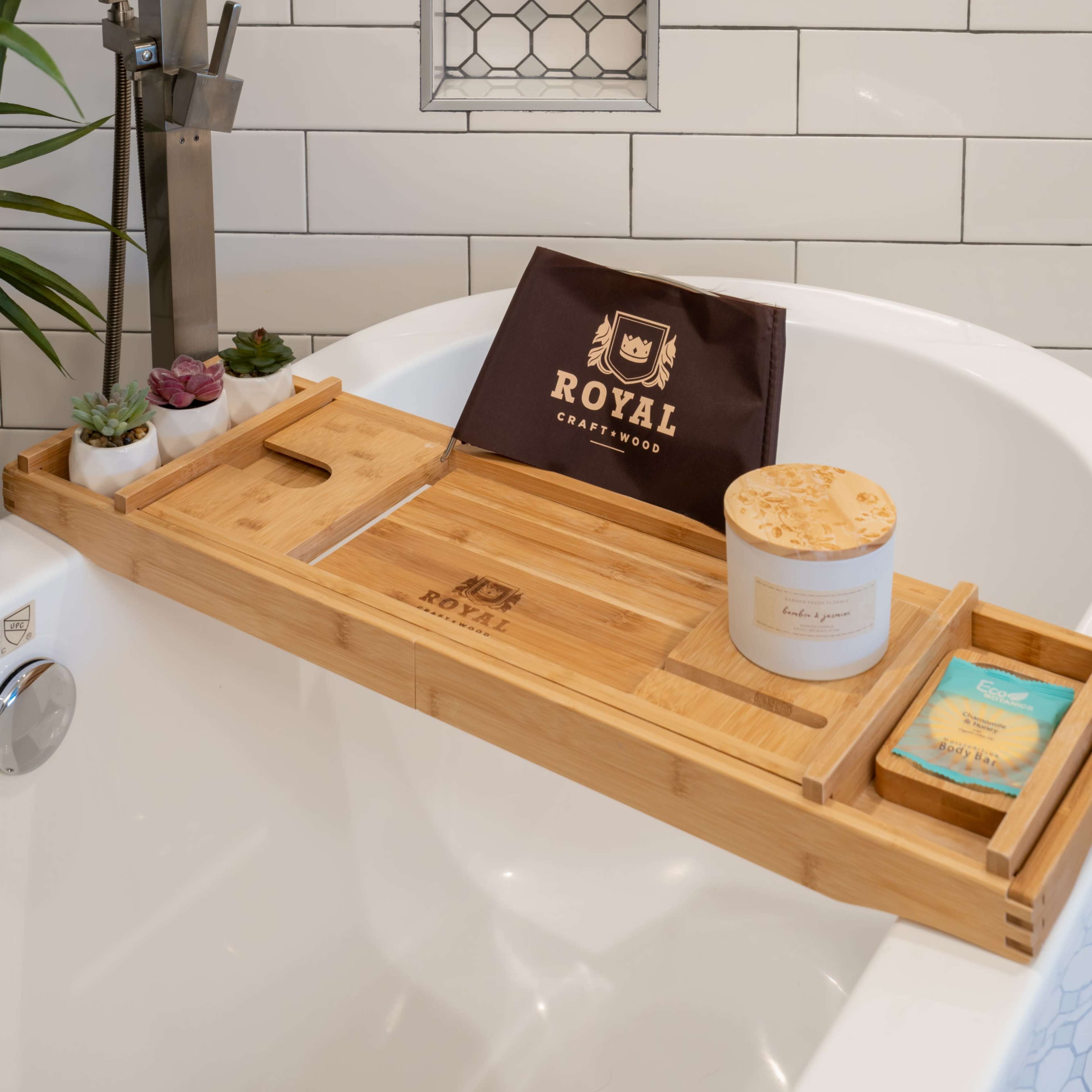 A bamboo bath tray rests across a white bathtub, displaying a candle, a bar of soap, and a small plant arrangement against a tiled wall.