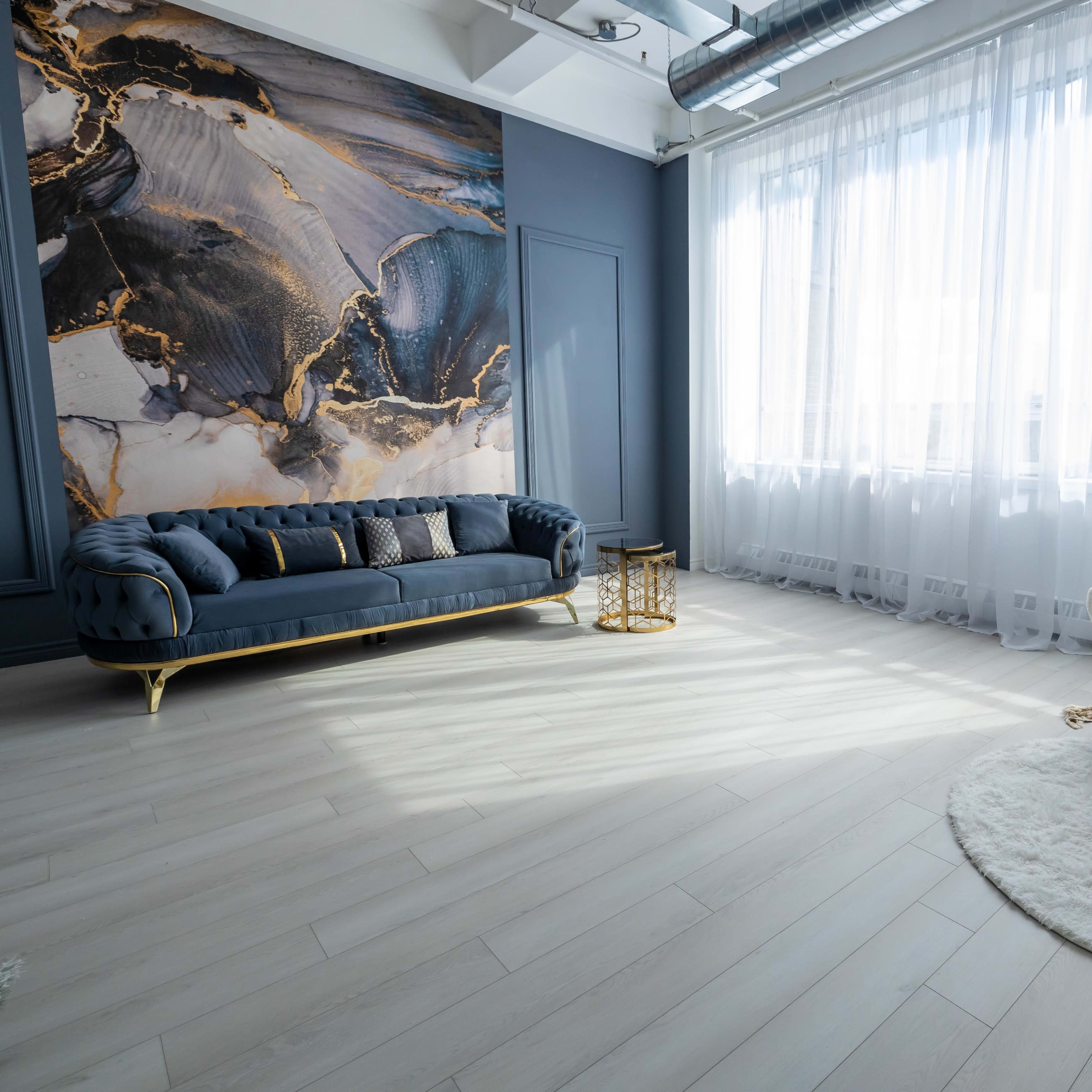 A well-lit room featuring a blue velvet sofa, a white circular rug, and a small wooden table, with large windows draped in sheer white curtains.