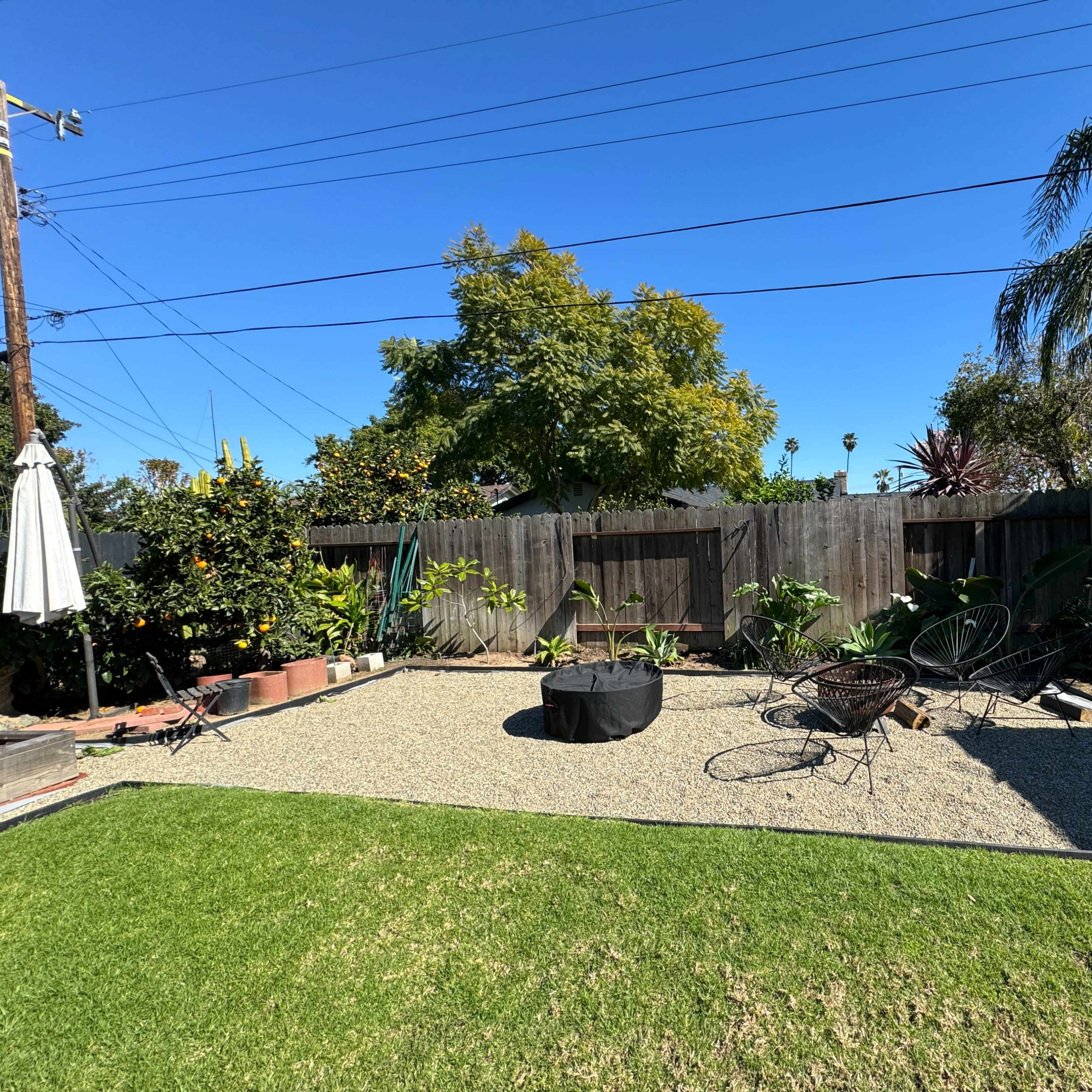 A landscaped backyard featuring a gravel area with circular seating, surrounded by plants and trees under a clear blue sky.