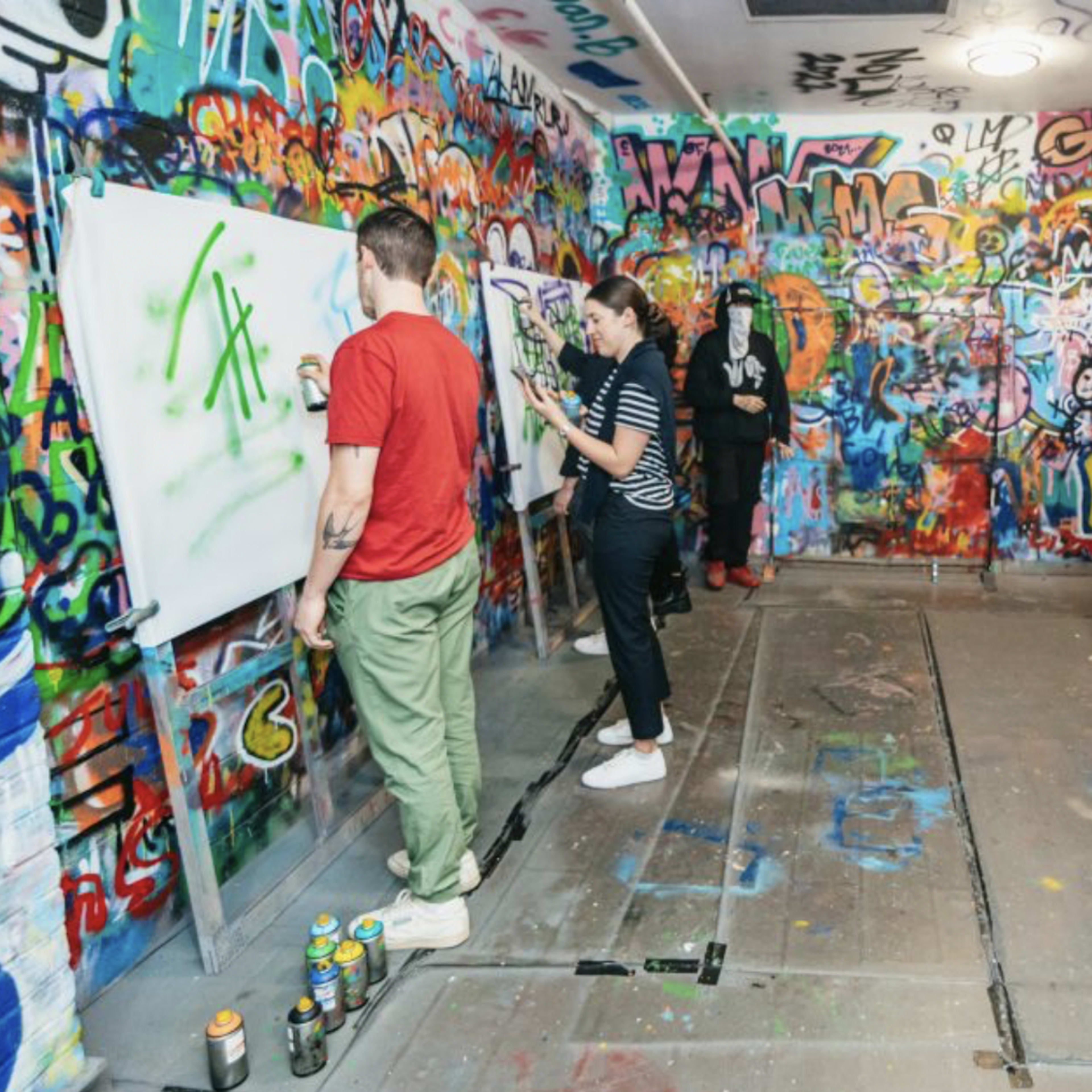 A group of people is engaged in painting graffiti on large white canvases in a brightly colored, graffiti-covered room.