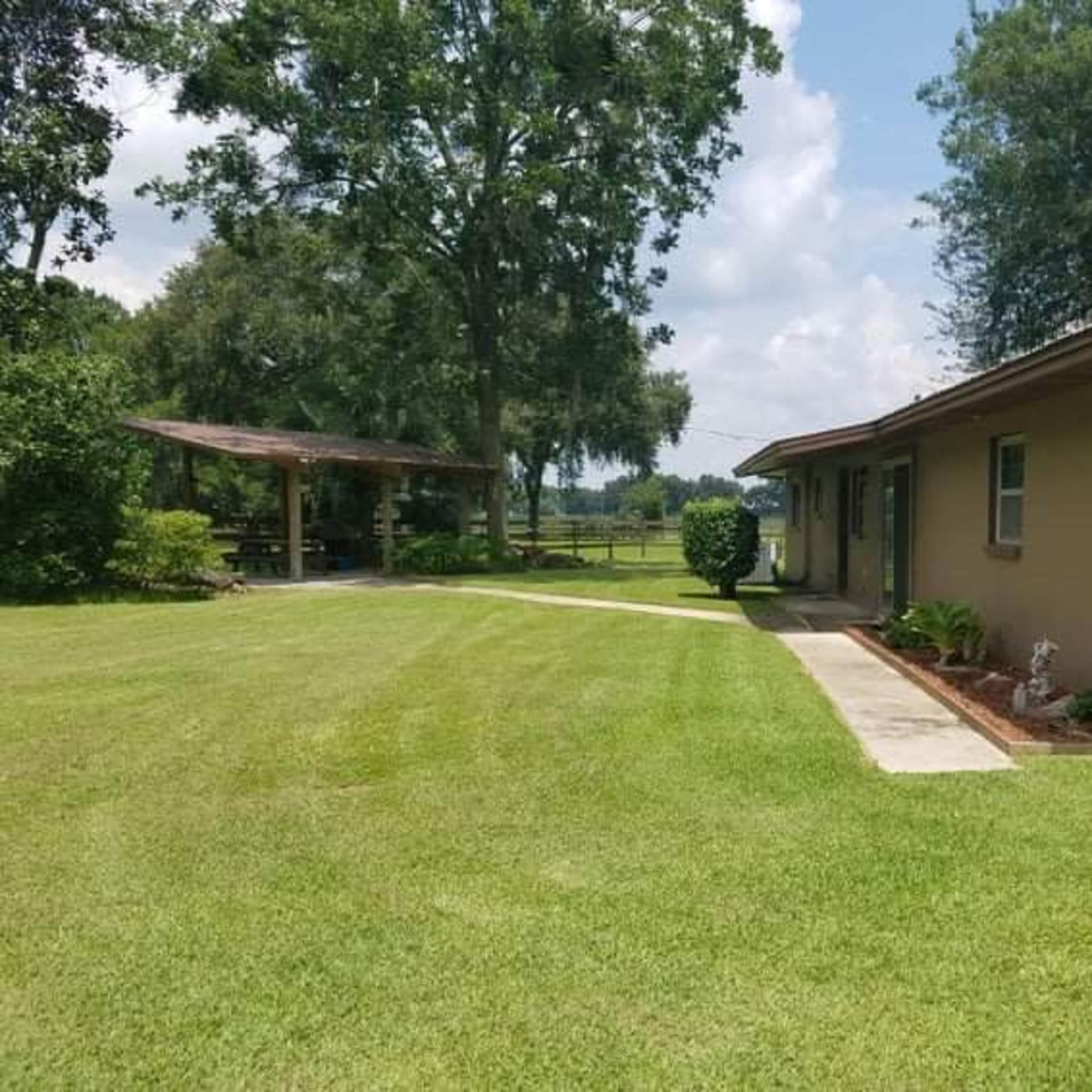A single-story house with a well-maintained lawn and a covered patio is visible, surrounded by trees and open fields.