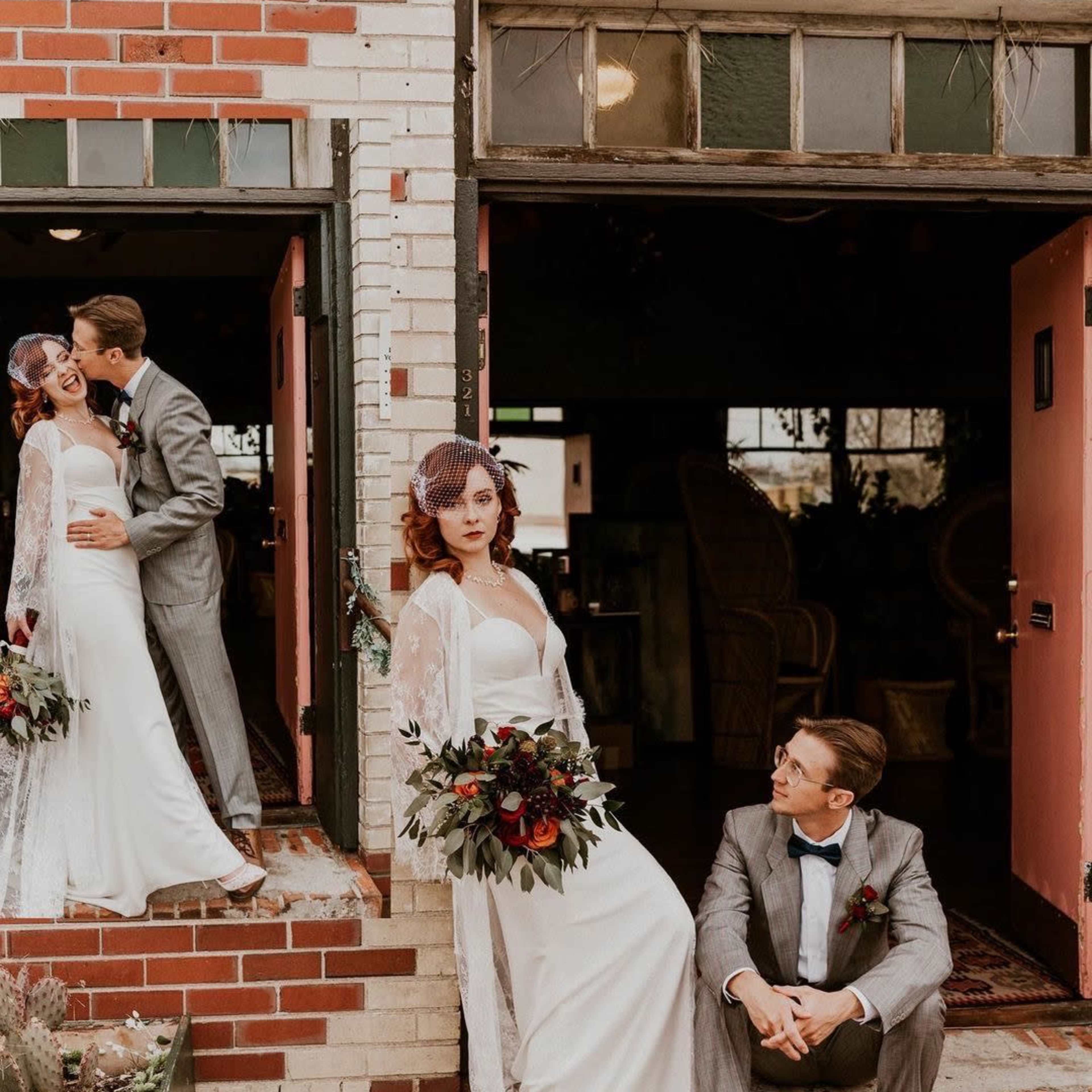 A bride in a white dress holds a bouquet while standing beside a groom in a gray suit, who is seated on the steps of a brick building.