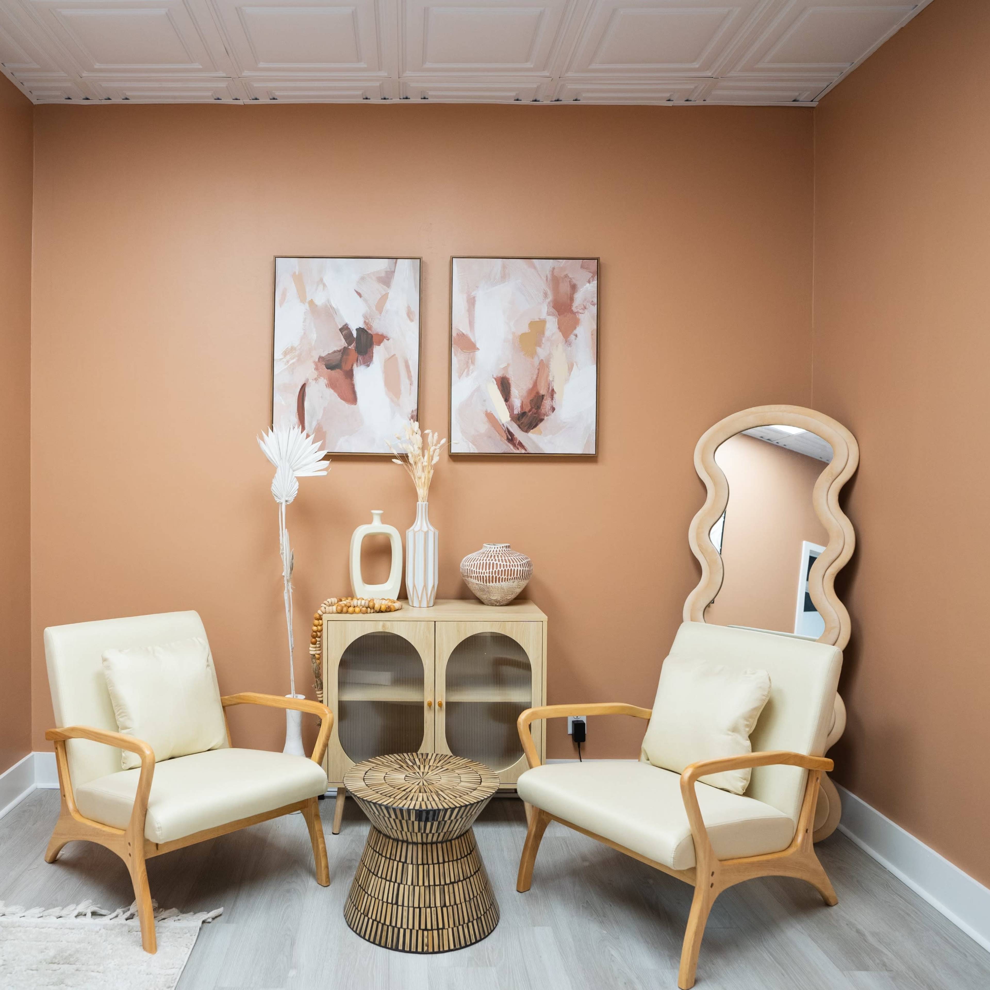 A small, stylish room with two light-colored chairs, a round table, decorative artwork on the walls, and a mirror reflecting the space.