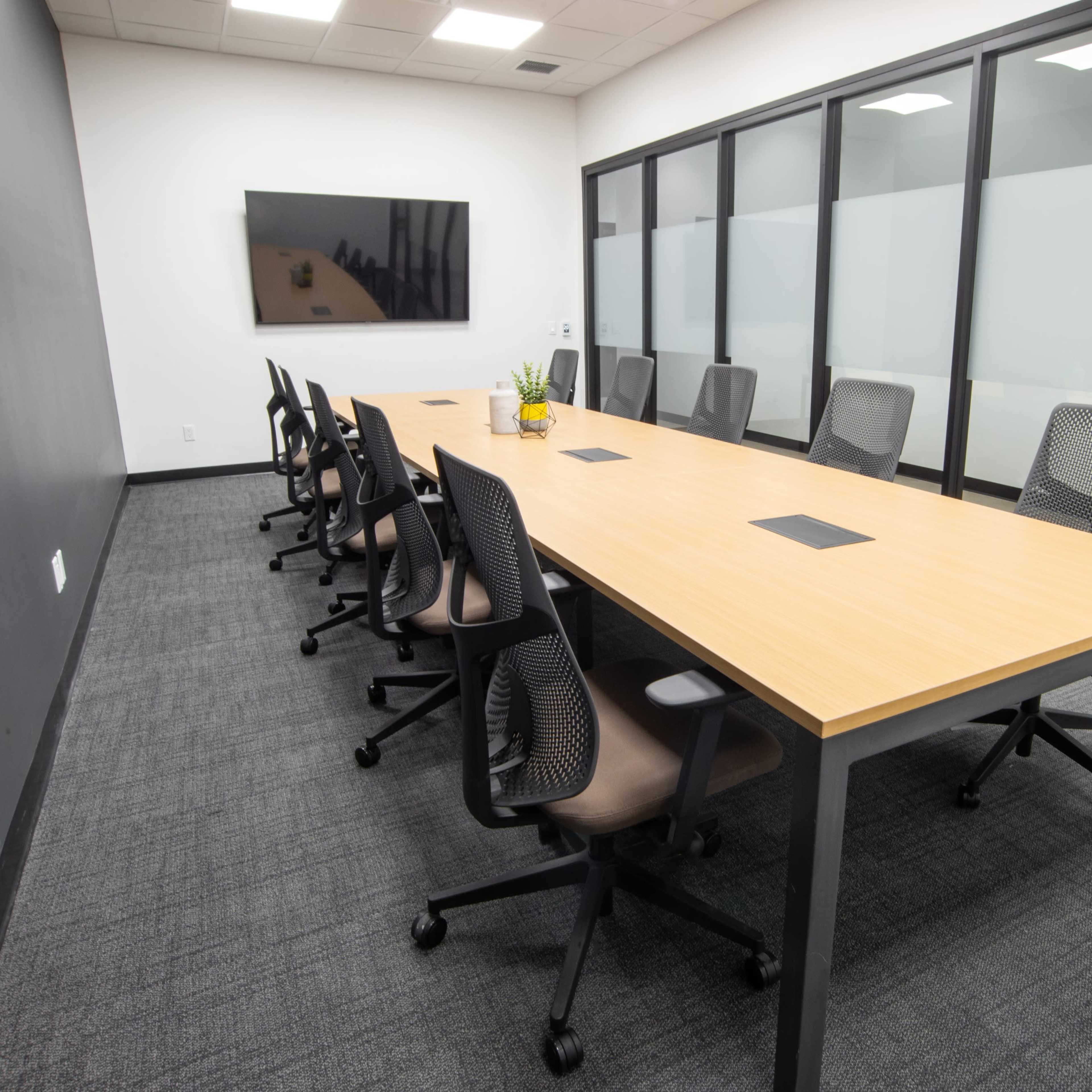 A modern conference room with a long wooden table surrounded by ergonomic chairs and a wall-mounted screen.