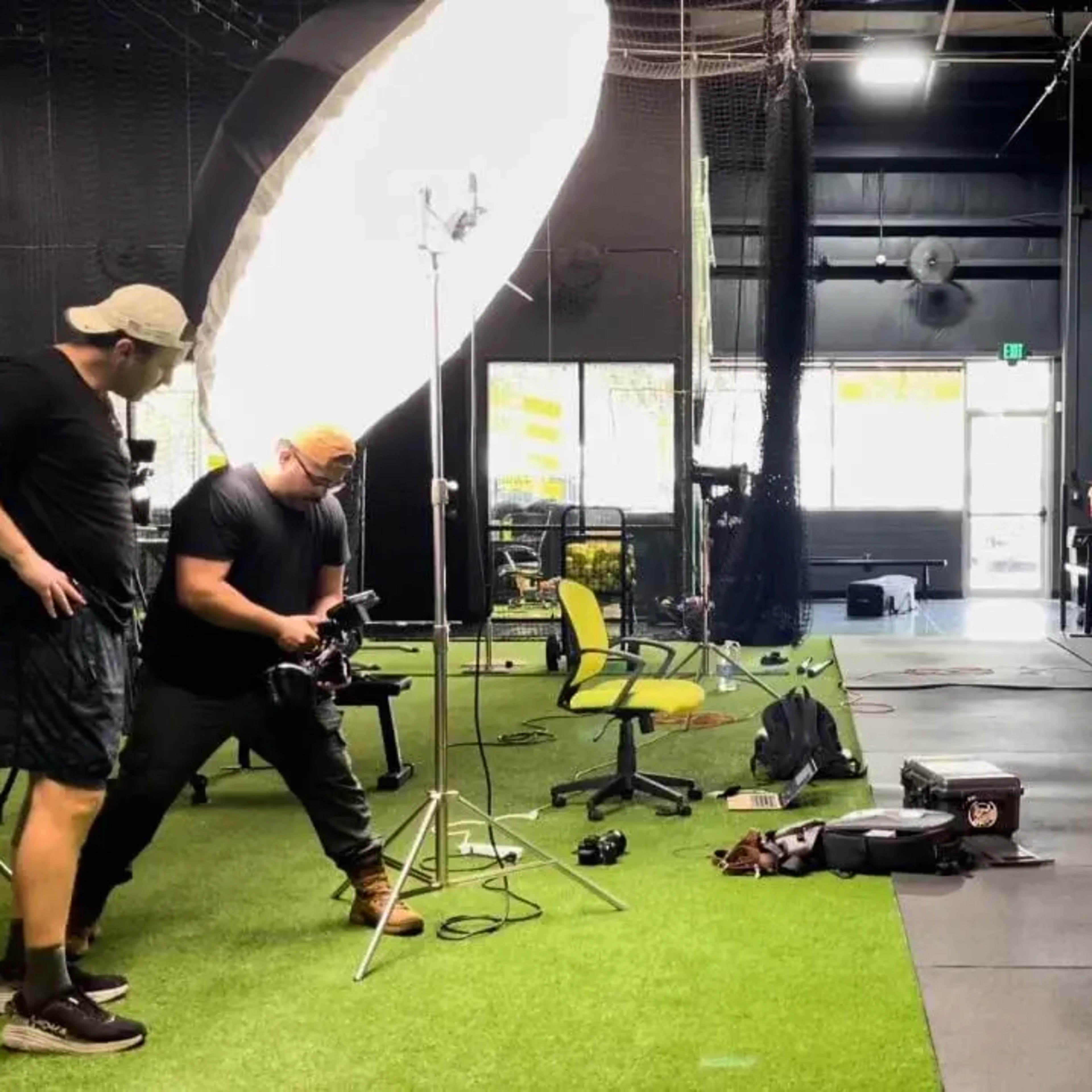 A photoshoot is taking place in a gym with a female model posing while two photographers adjust their equipment.