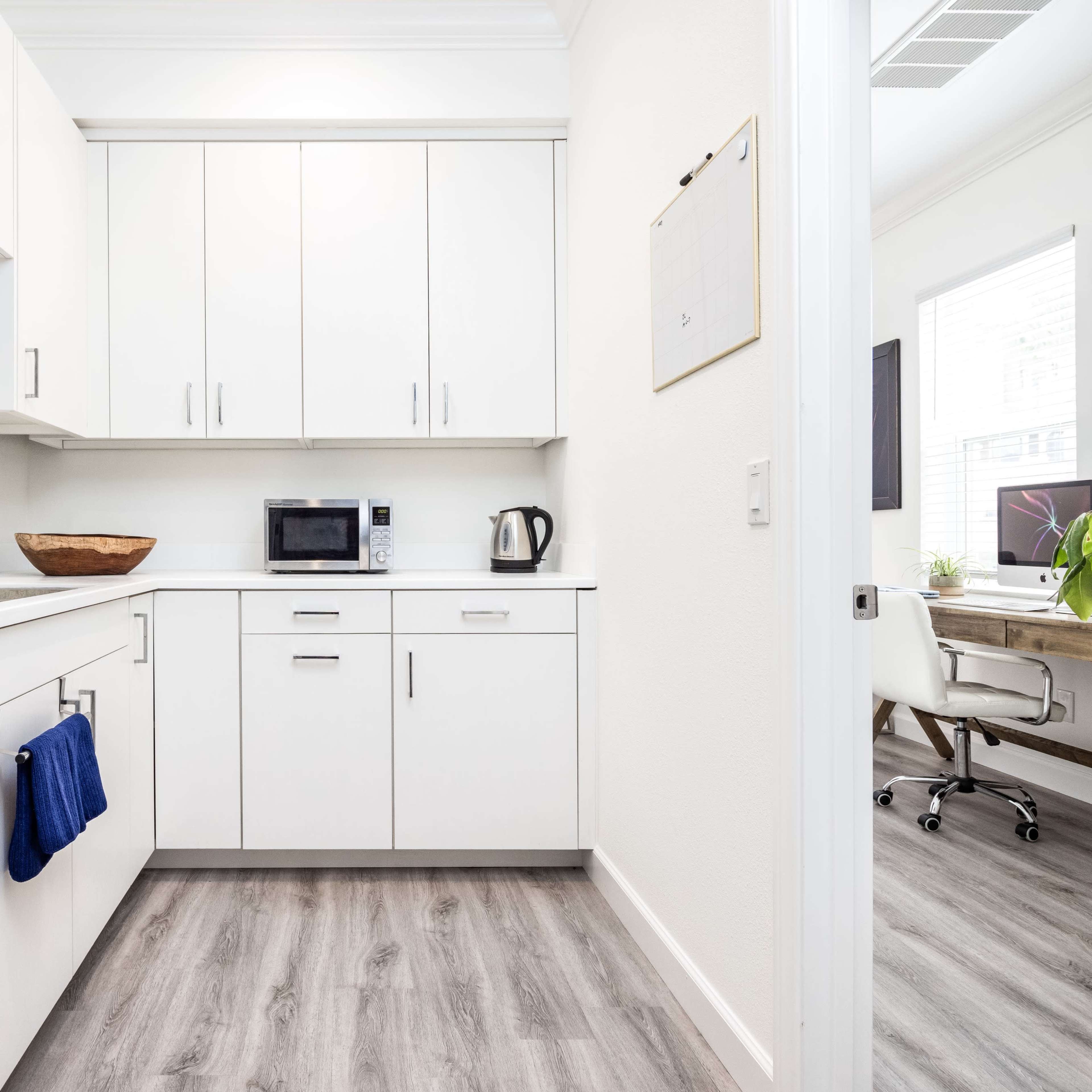A modern kitchen features white cabinetry, a sink, a microwave, and a kettle against a light-colored wall.