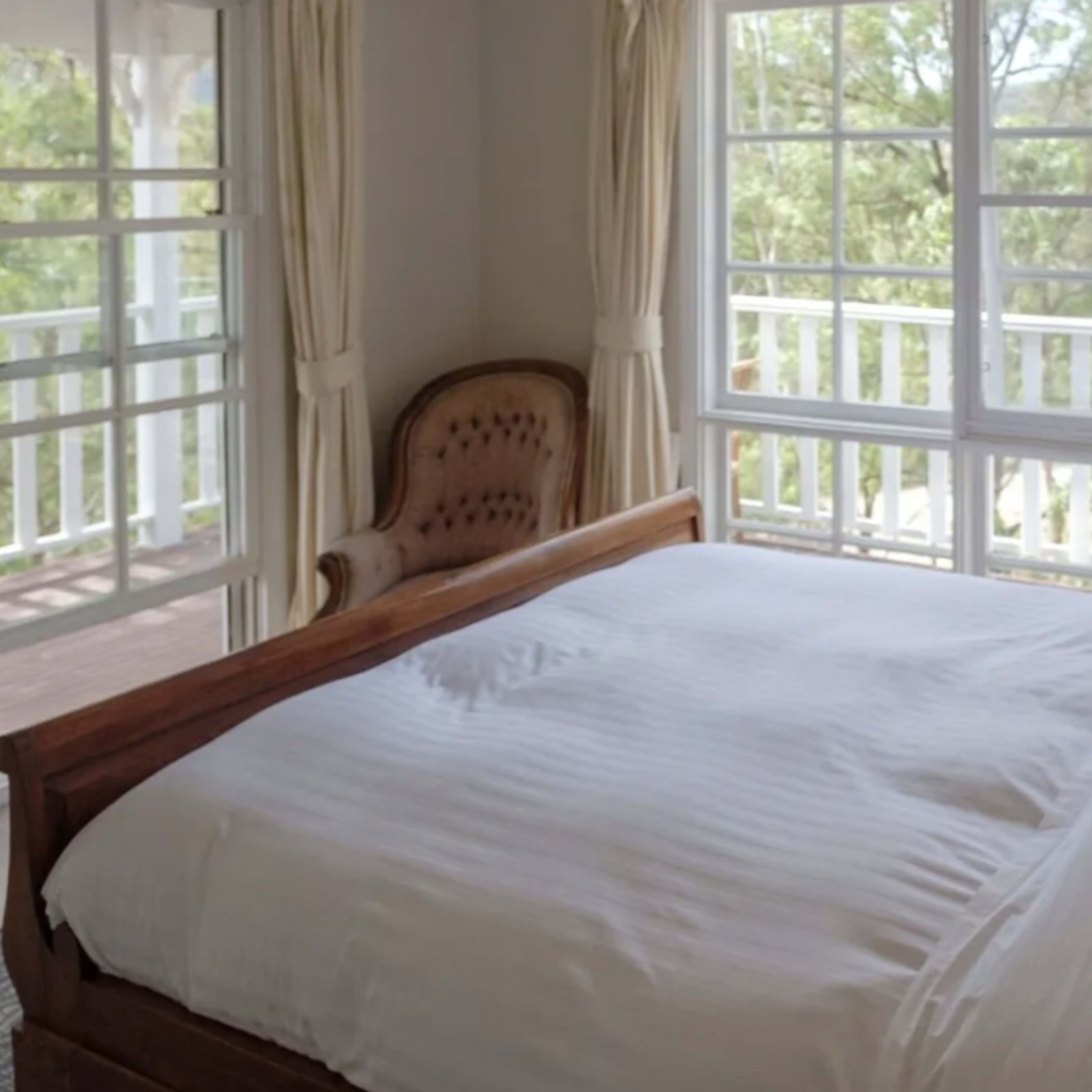 A bright bedroom features a wooden bed with white bedding, a chair in the corner, and large windows overlooking greenery.