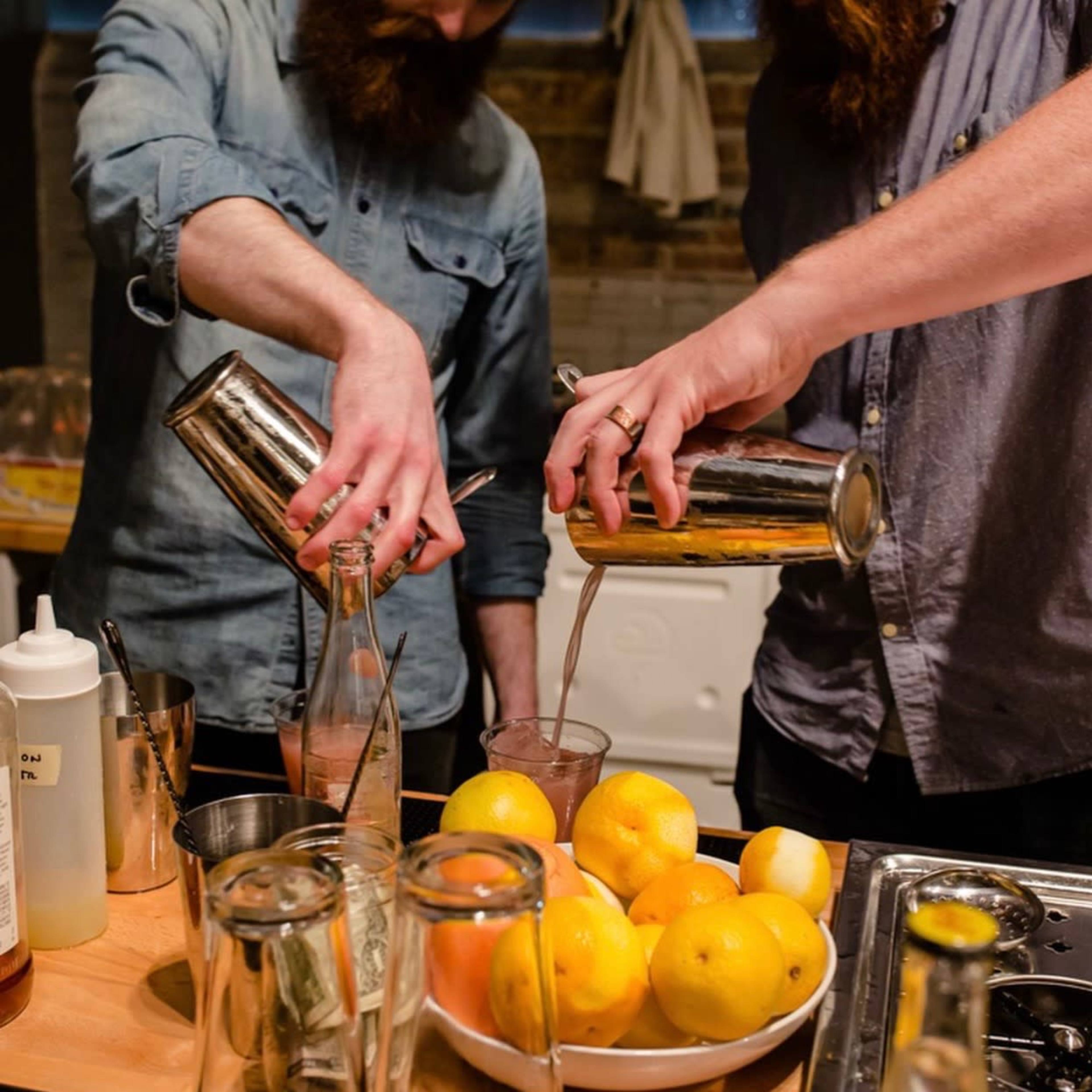 Two individuals with beards are pouring drinks from metal shakers into glasses while surrounded by bottles and a bowl of lemons on a kitchen counter.