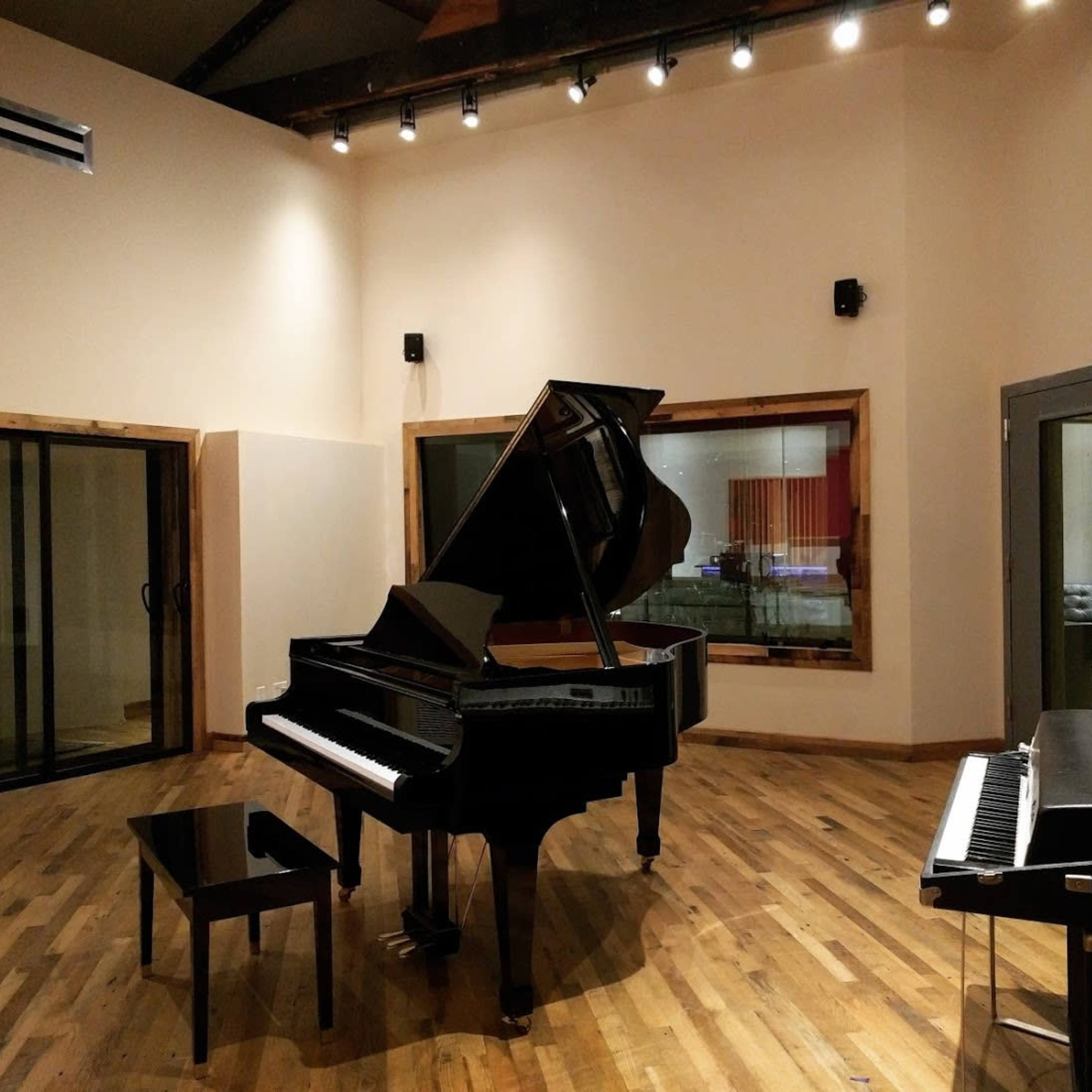 A grand piano and a small black upright piano are positioned in a spacious recording studio with wooden floors and large glass doors.