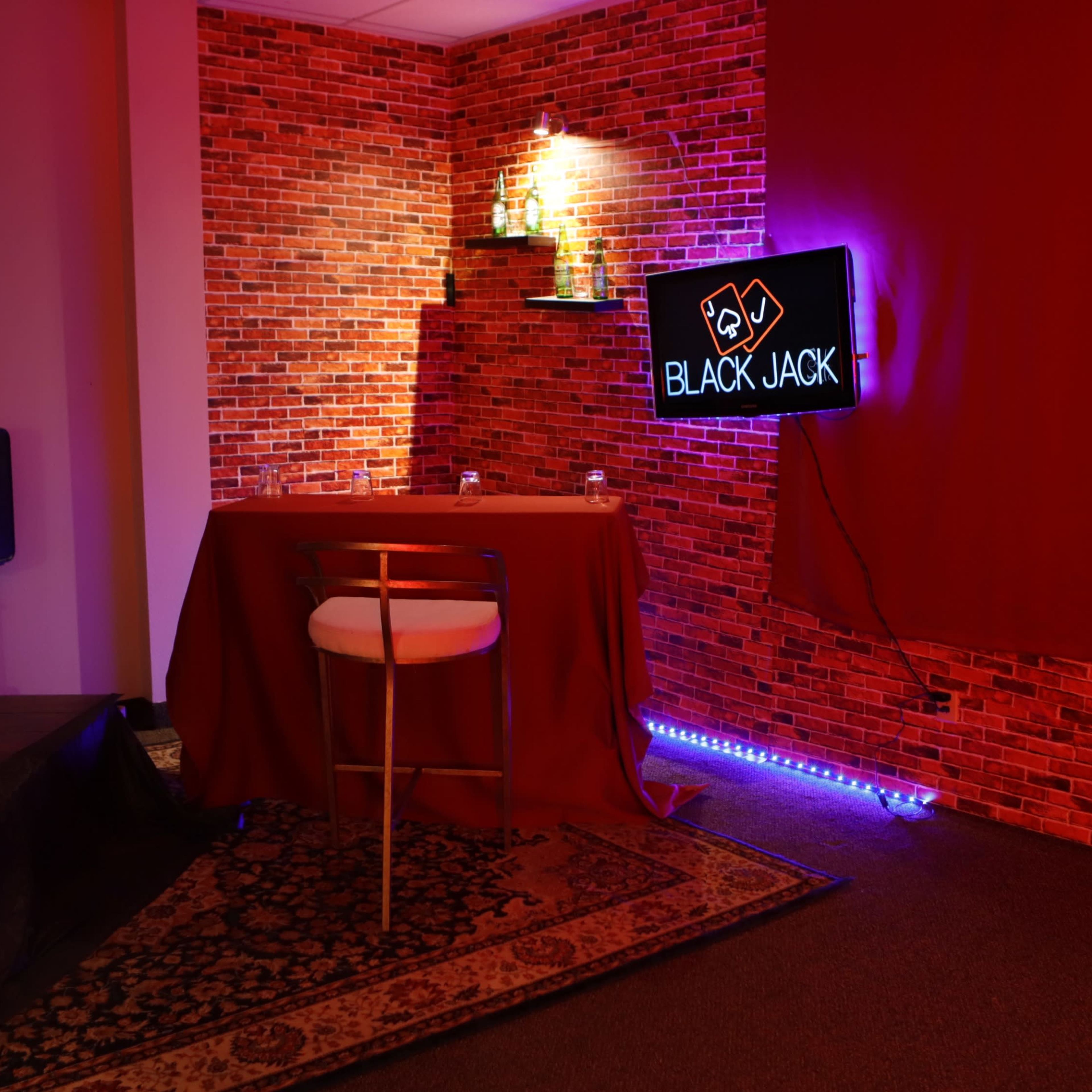 A small room features a red-brick wall, a neon "BLACKJACK" sign, and a single table with a white chair, illuminated by ambient lighting.
