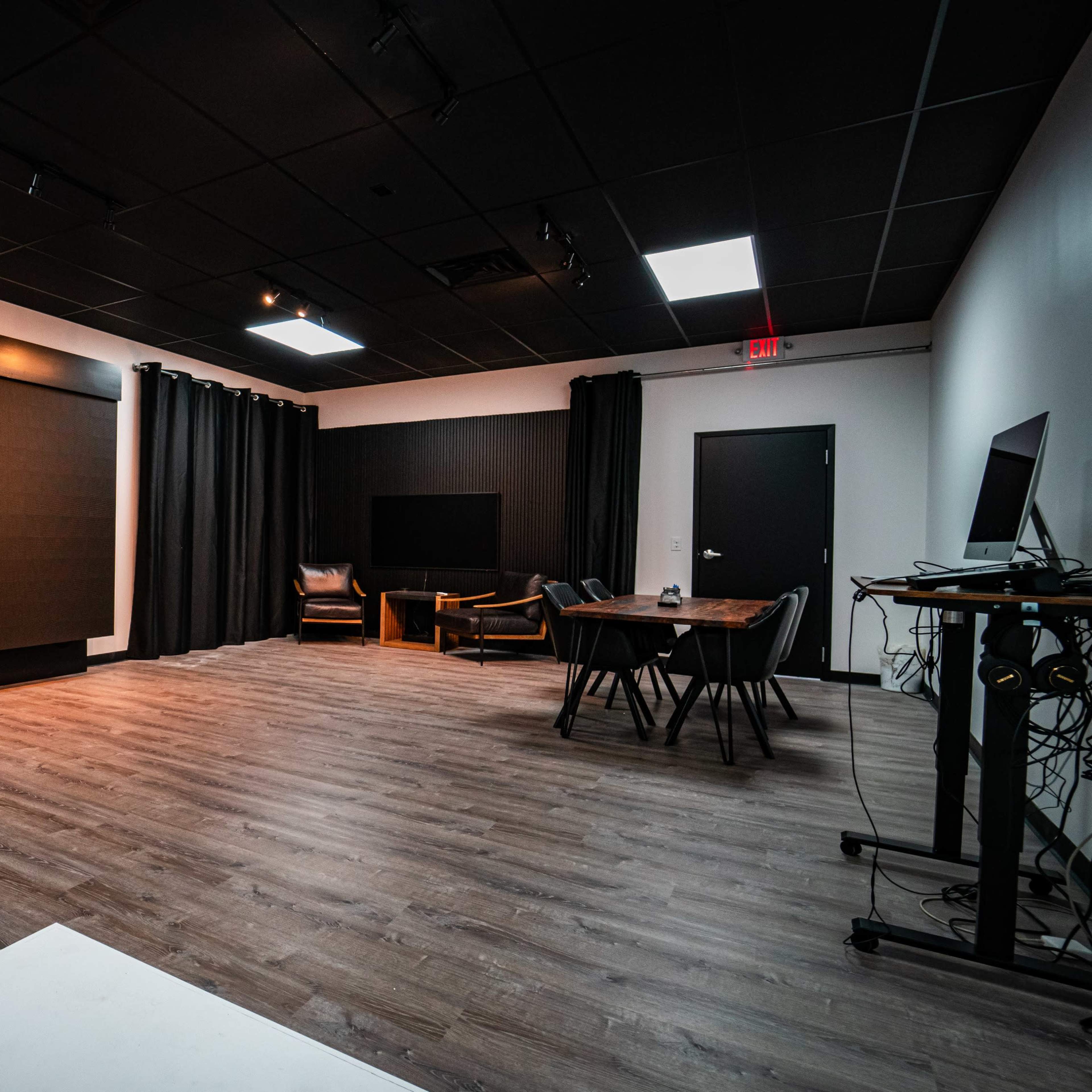 A large, empty room features a dark wall-mounted screen, a table with chairs, and black curtains along the walls.