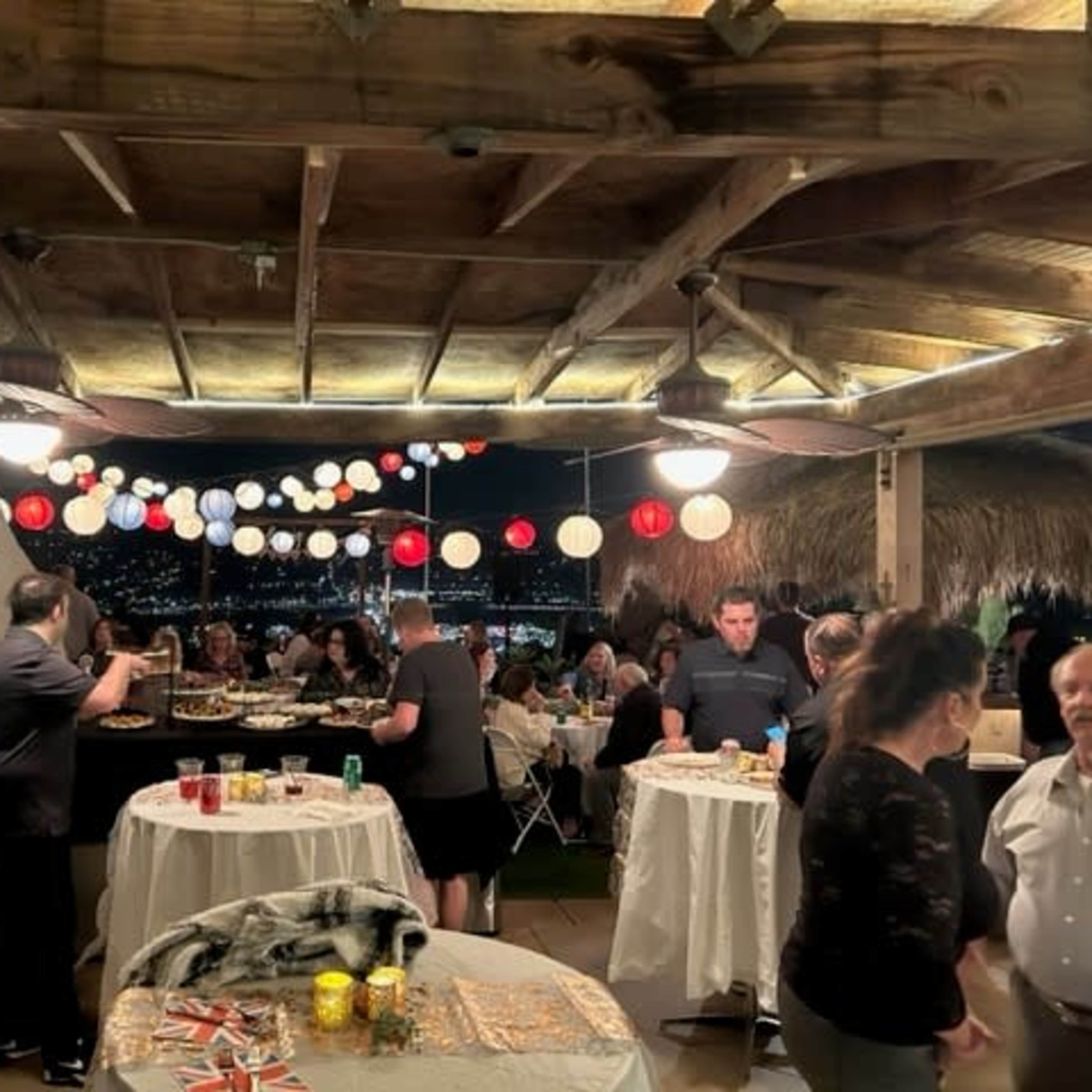 A group of people gathers under a wooden roof adorned with colorful lanterns, enjoying food and drinks at a festive outdoor event.