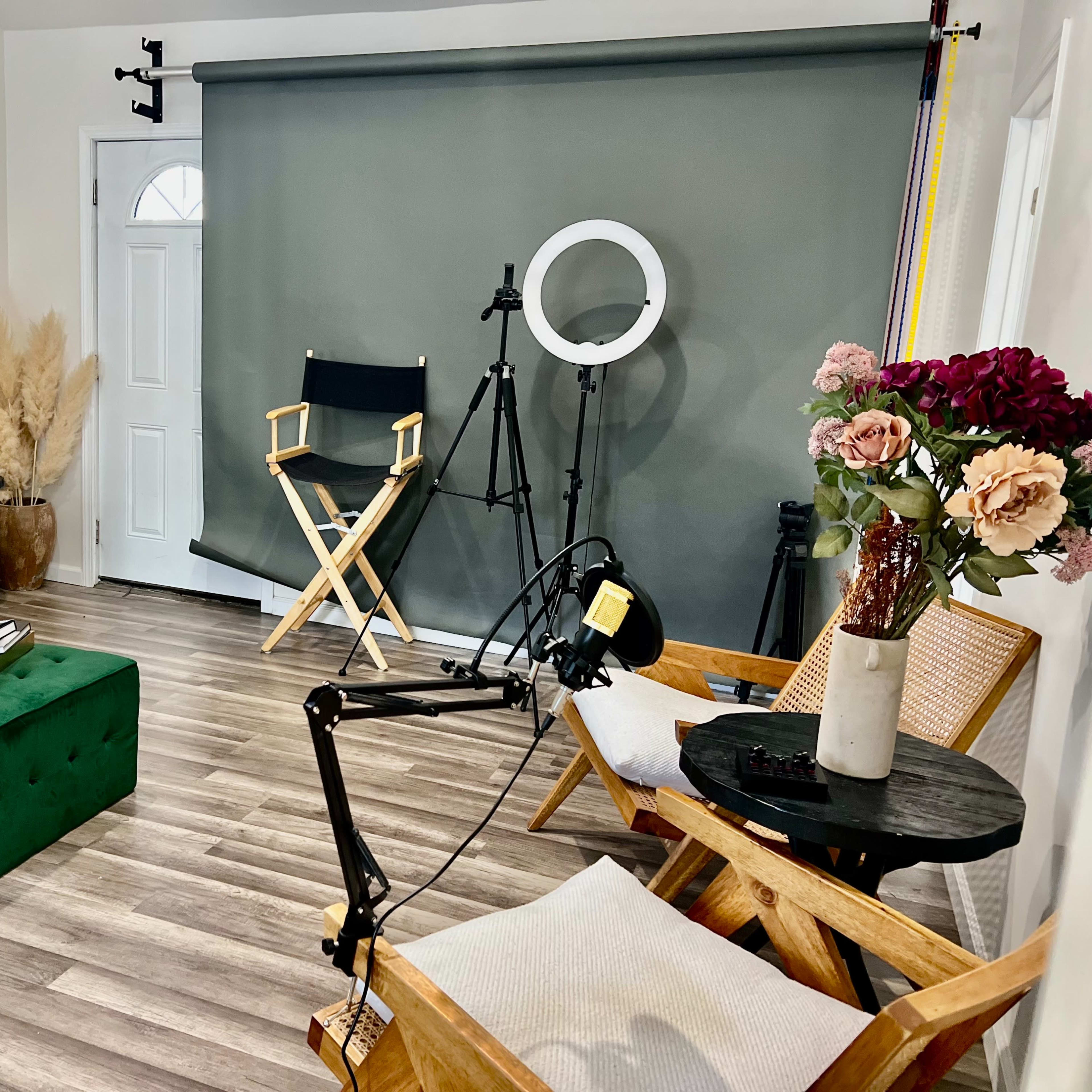 A well-decorated room featuring a green velvet sofa, a matching ottoman, a director's chair, a ring light, and potted flowers, all arranged around a wooden table.