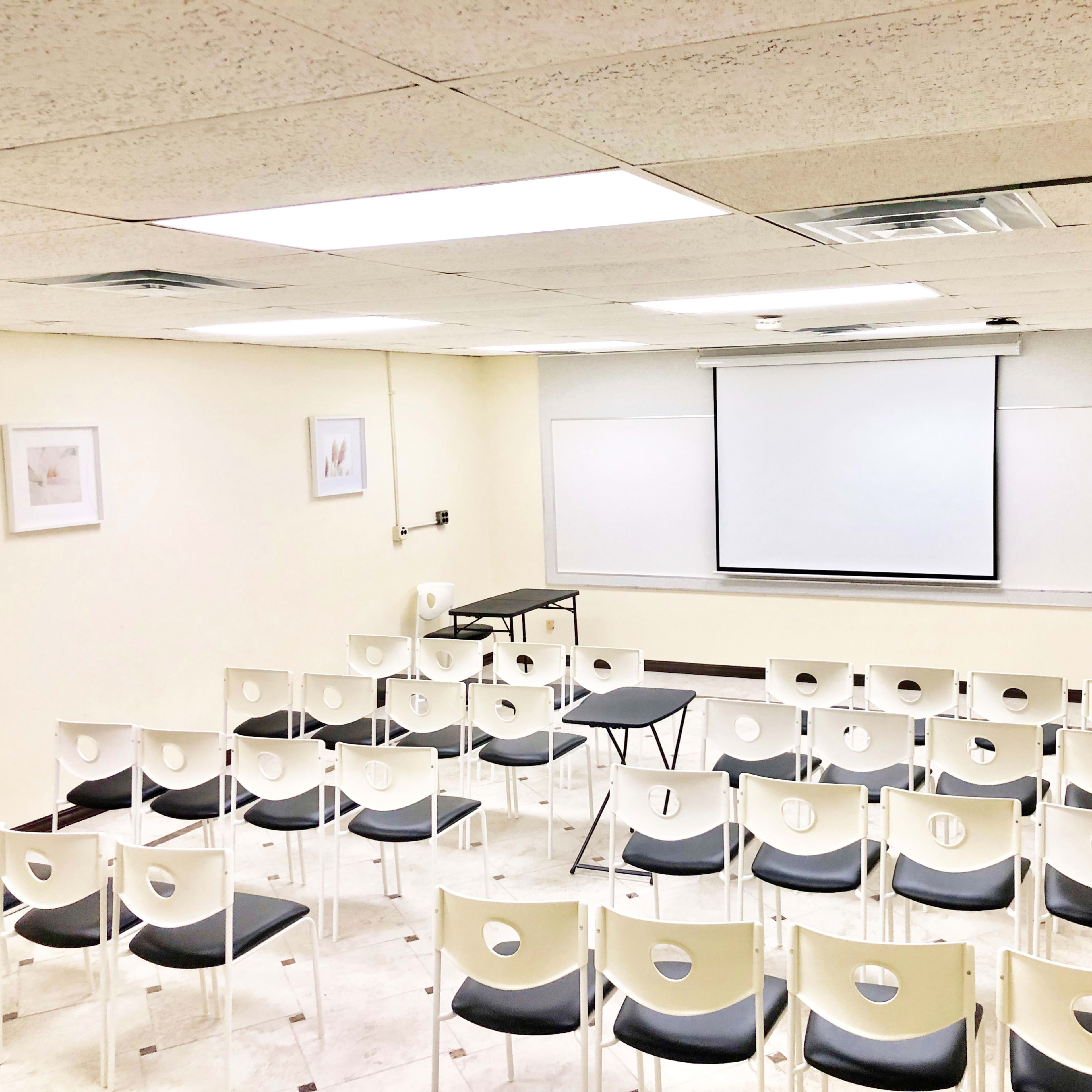 A classroom is arranged with rows of white chairs facing a projector screen at the front.