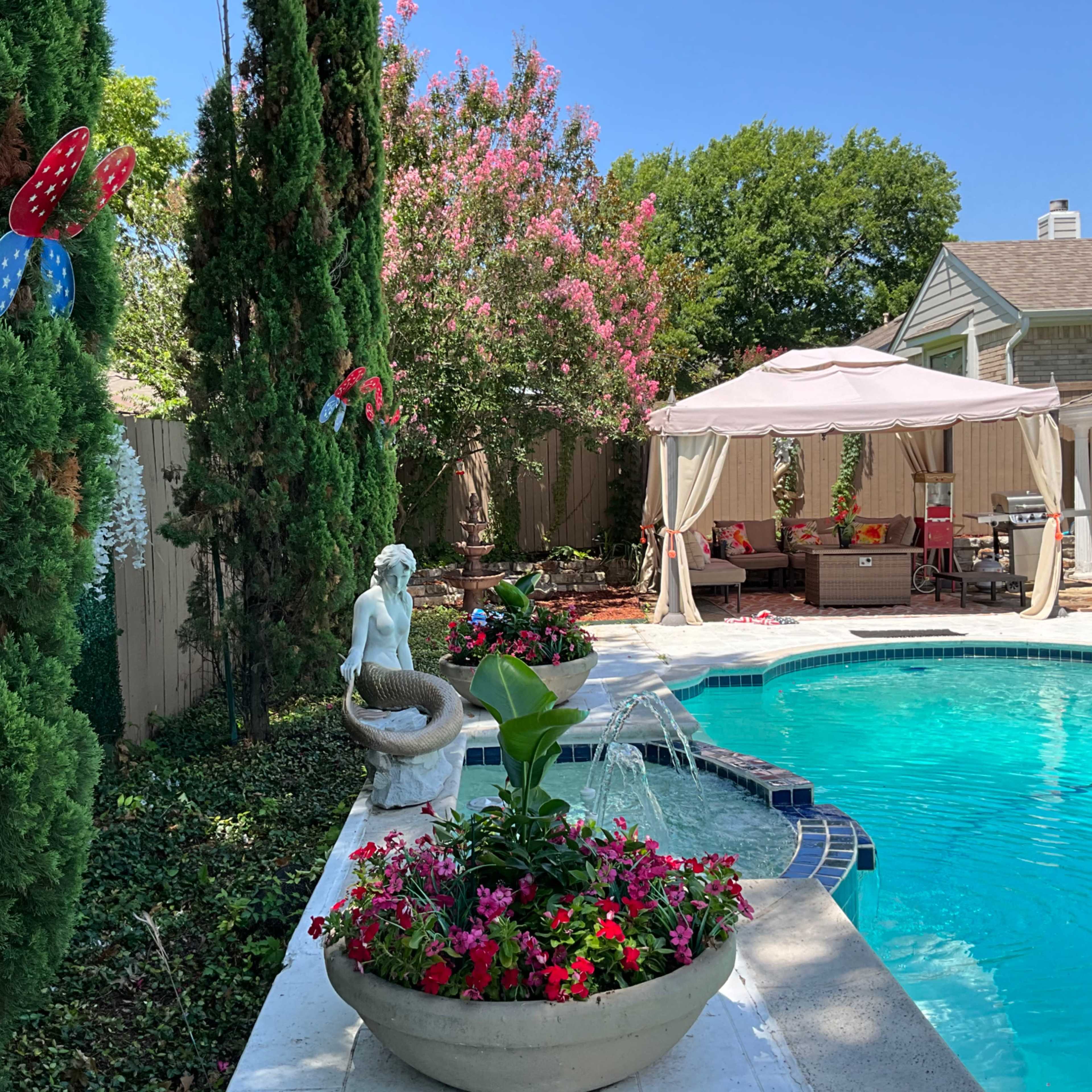 A swimming pool surrounded by colorful flower beds, a statue, and a shaded seating area with a canopy.