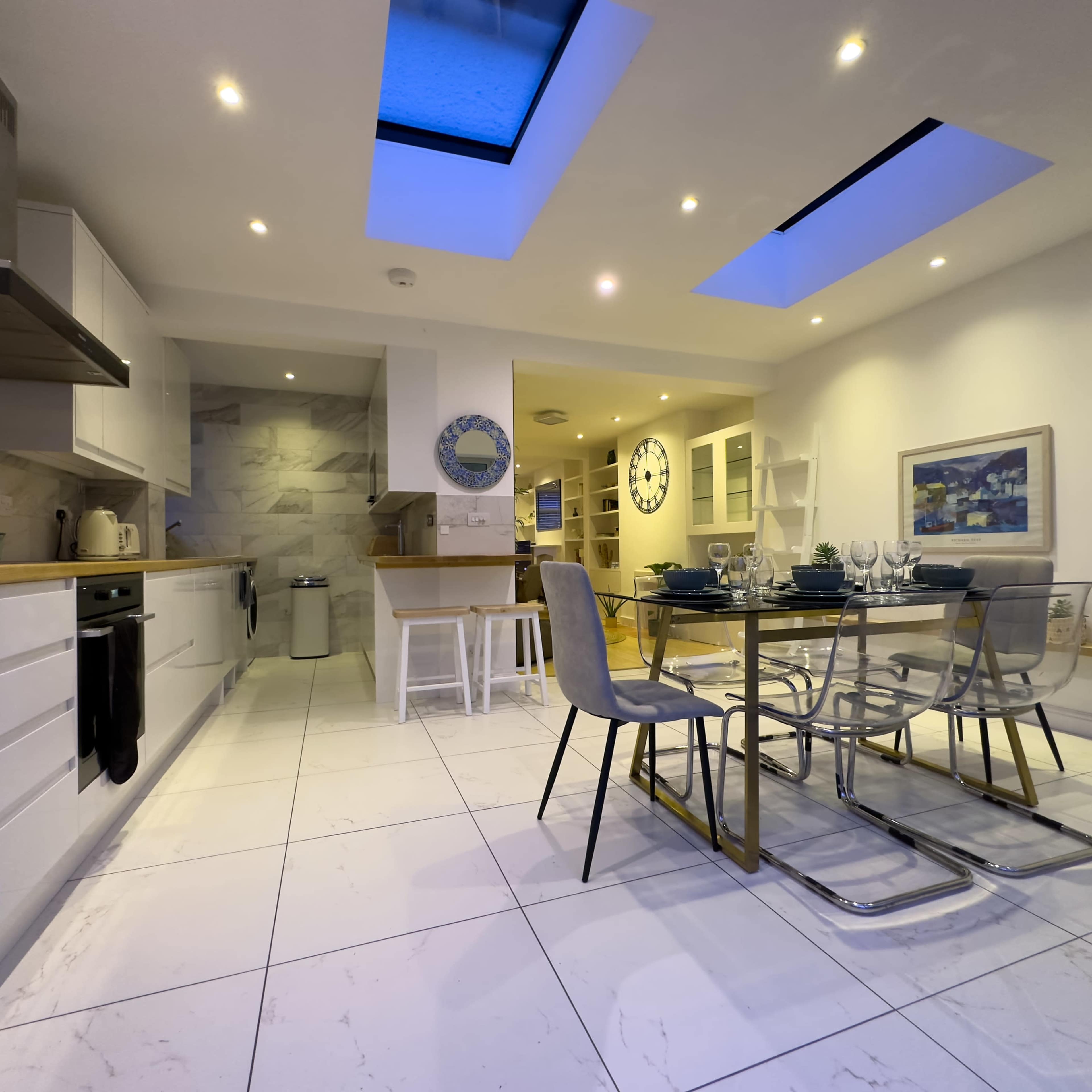 A modern kitchen features a dining table set for six, with light-colored cabinetry and skylights providing natural illumination.
