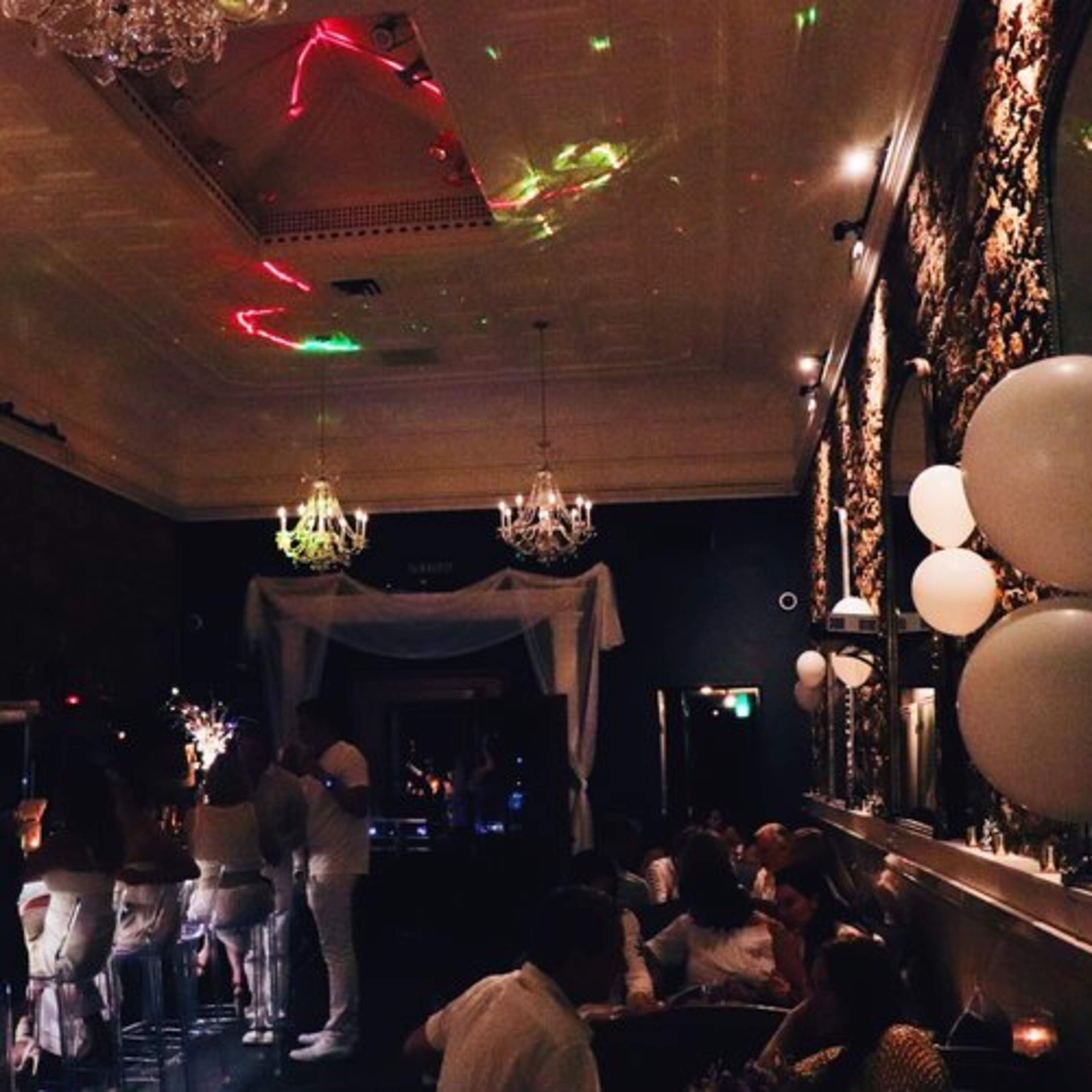 A dimly lit bar features people mingling while balloons and colorful lights illuminate the ceiling.