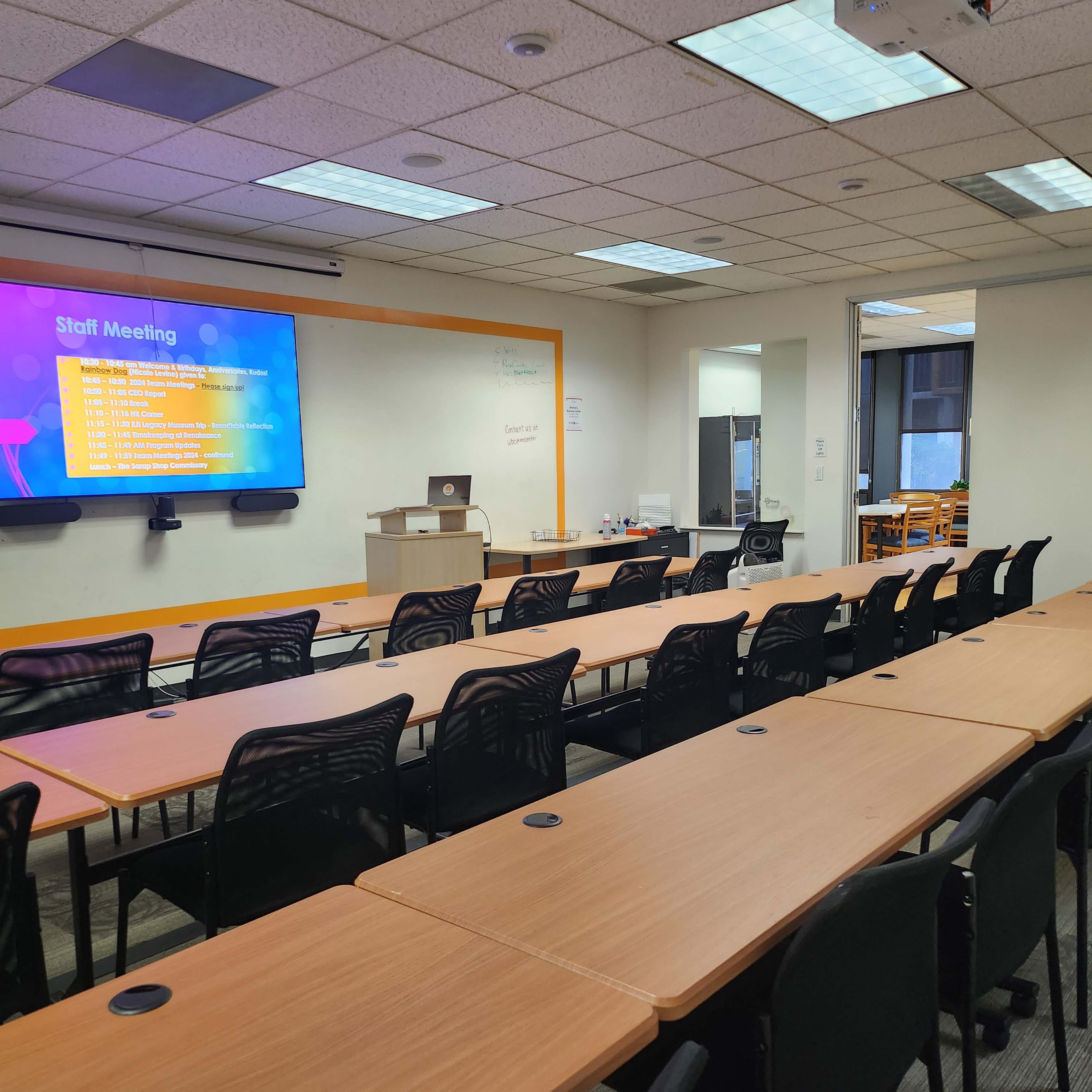 A conference room features rows of tables and chairs, with a presentation screen displaying meeting agenda items at the front.