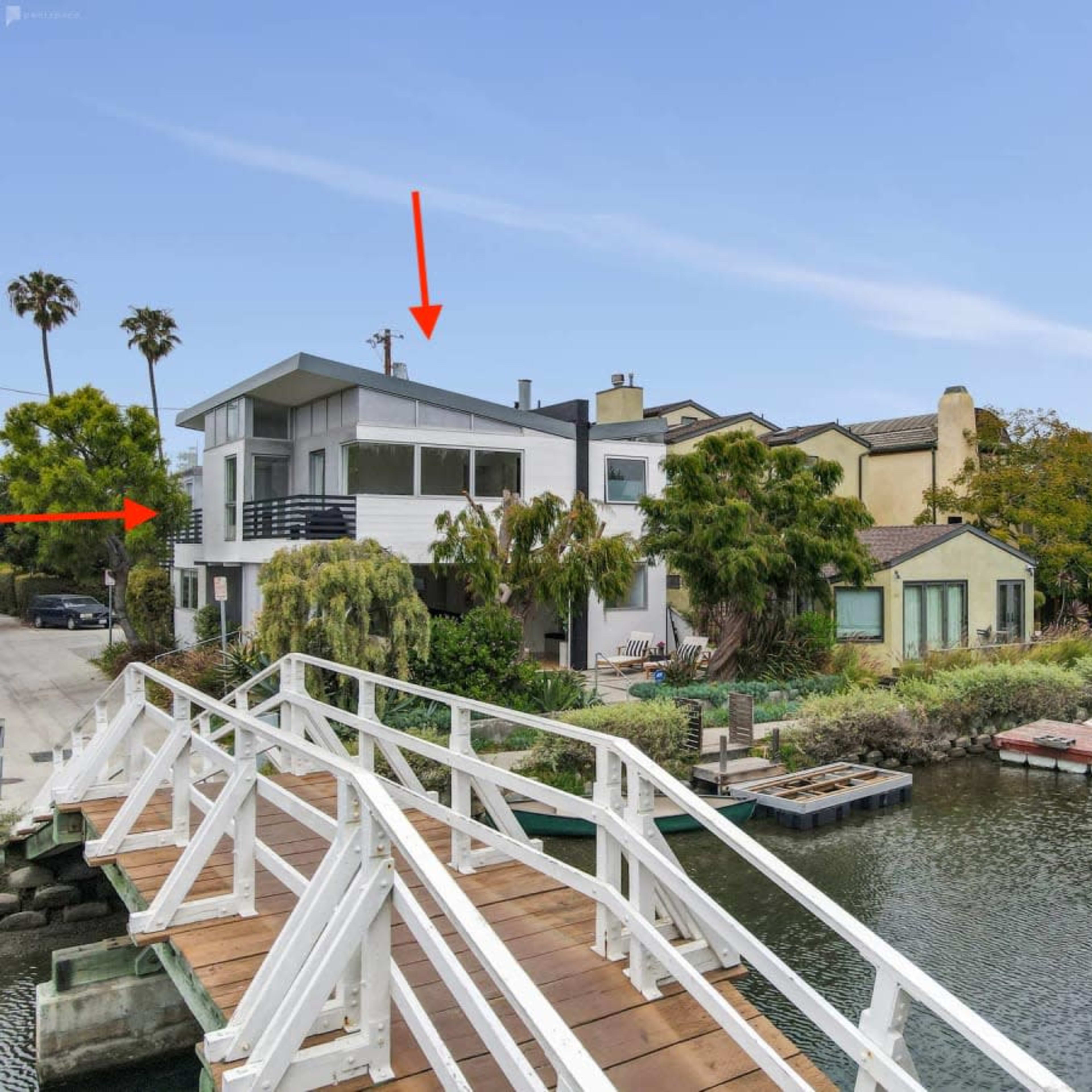A modern two-story house with large windows overlooks a calm waterway, connected by a wooden bridge to a street lined with palm trees.