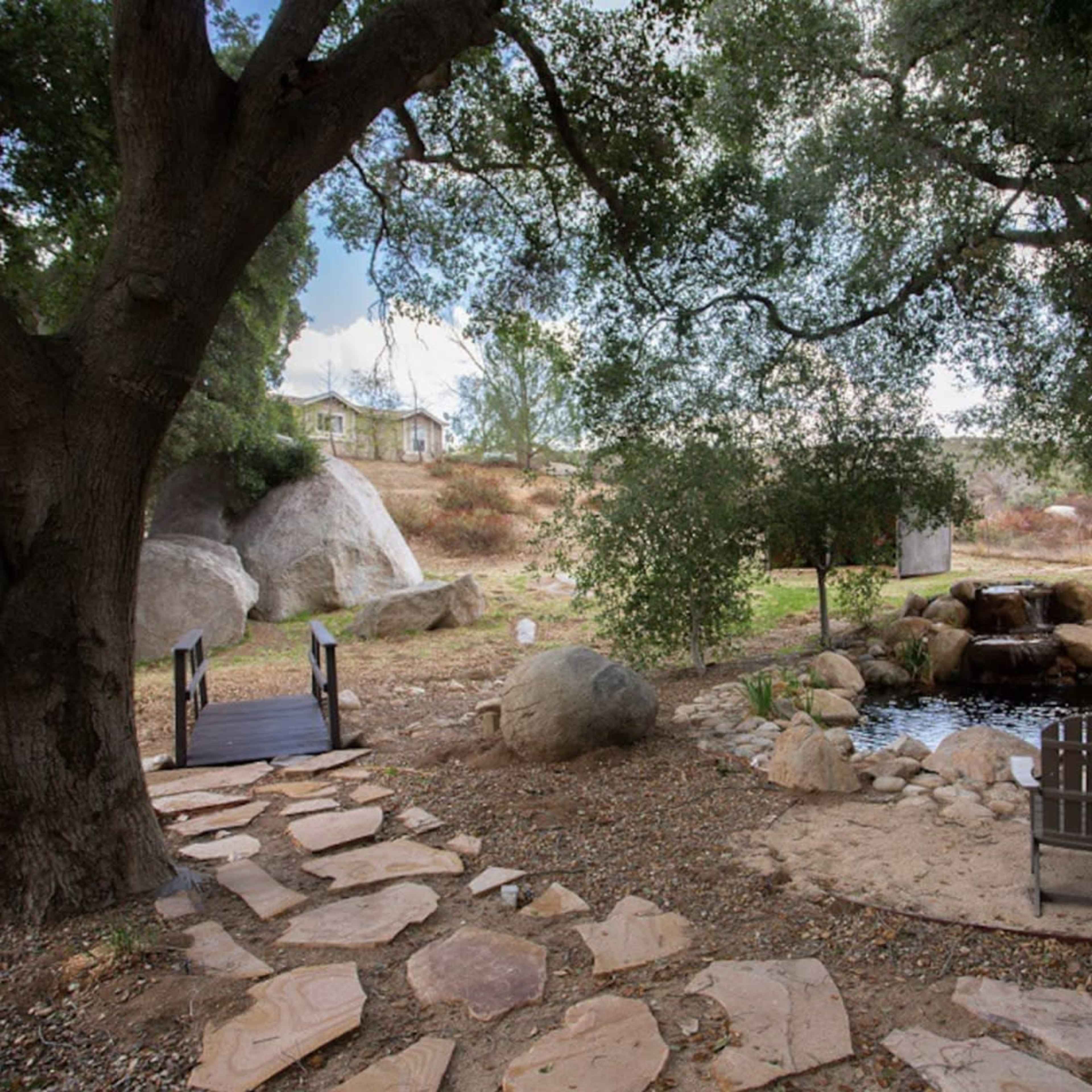 A stone path leads to a pond surrounded by large rocks and two wooden chairs.