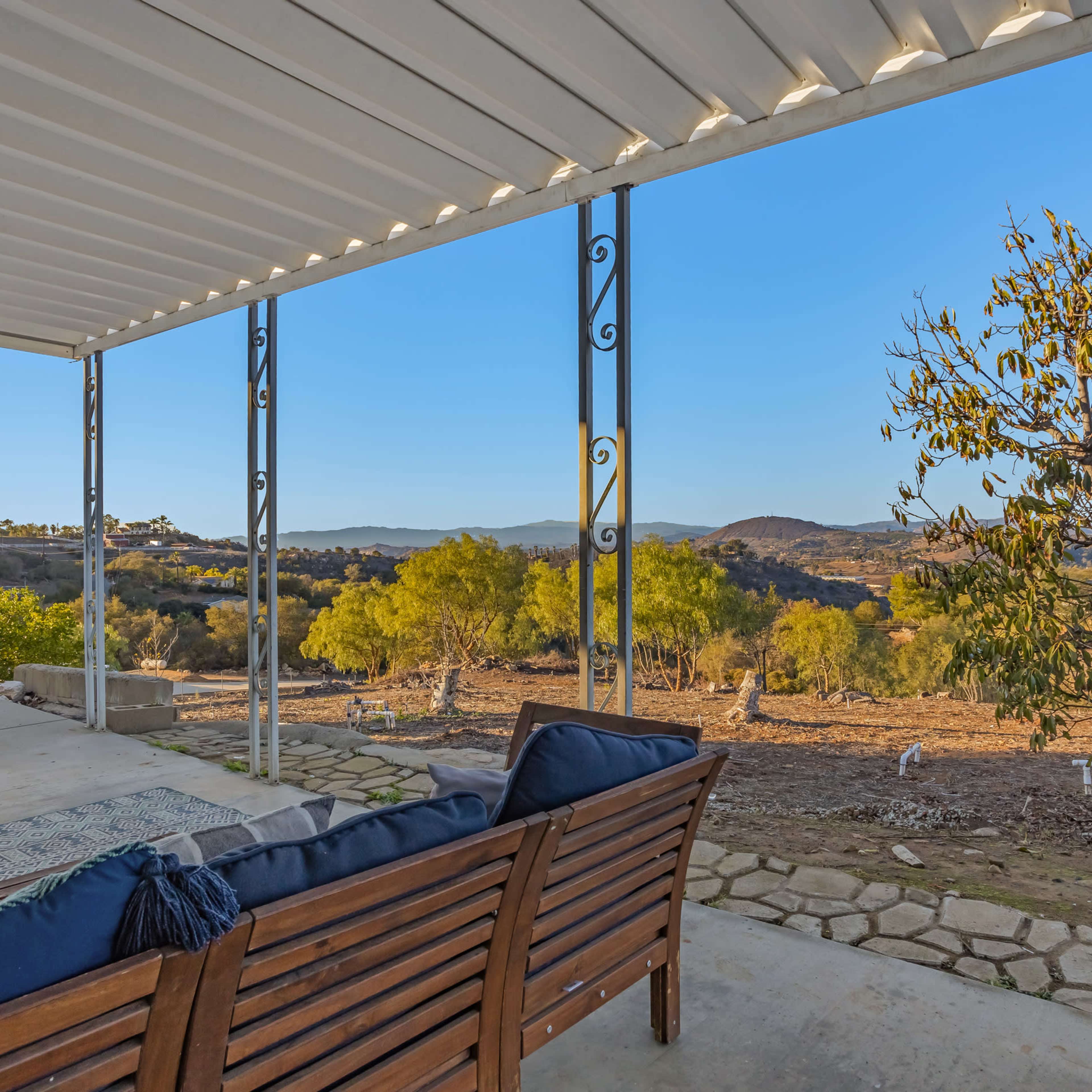 A covered patio with wooden seating overlooks a hilly landscape filled with trees under a clear blue sky.
