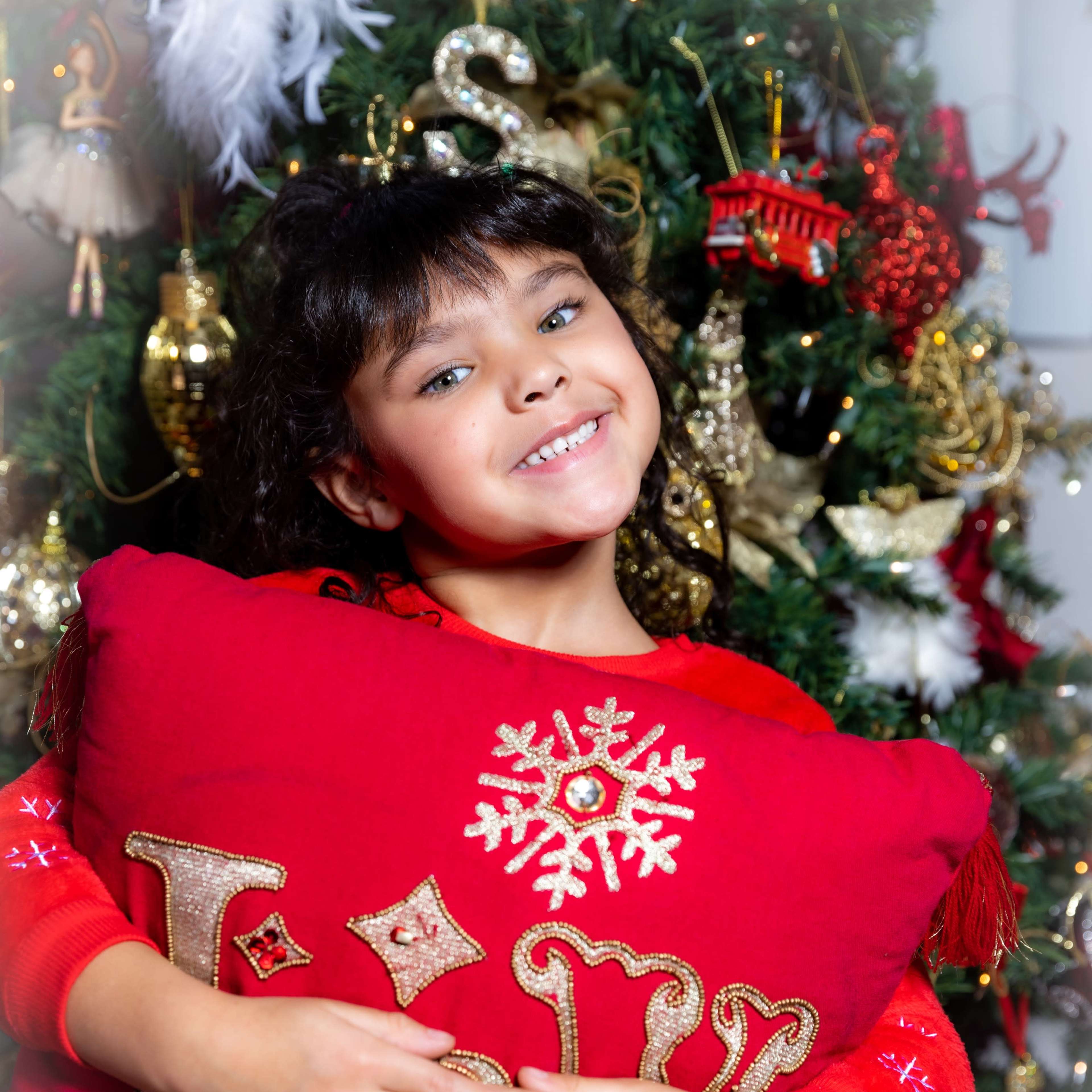 A girl smiles while holding a red decorative pillow in front of a Christmas tree adorned with ornaments and lights.