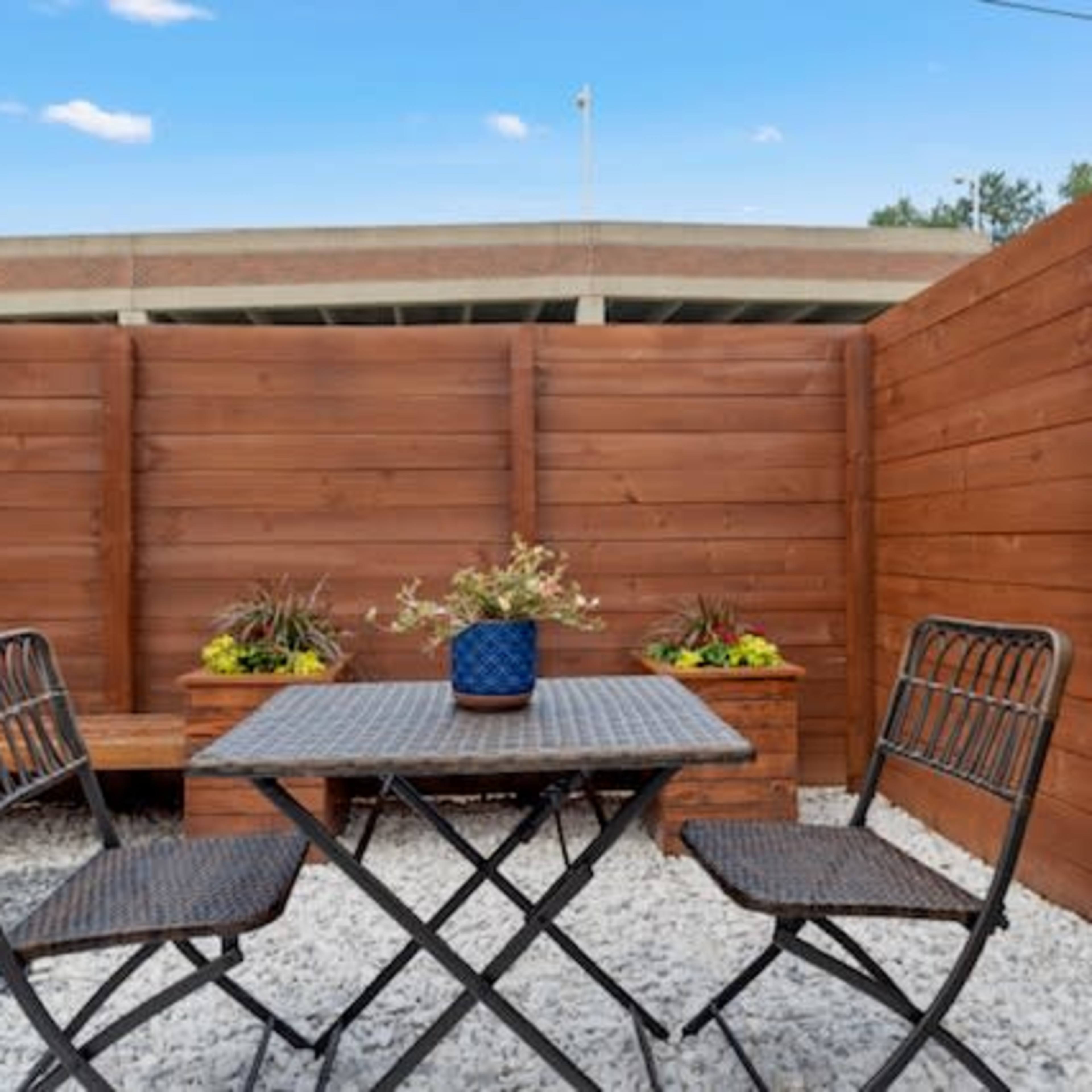 A small outdoor seating area with a metal table and two chairs surrounded by wooden fences and planters filled with flowers.