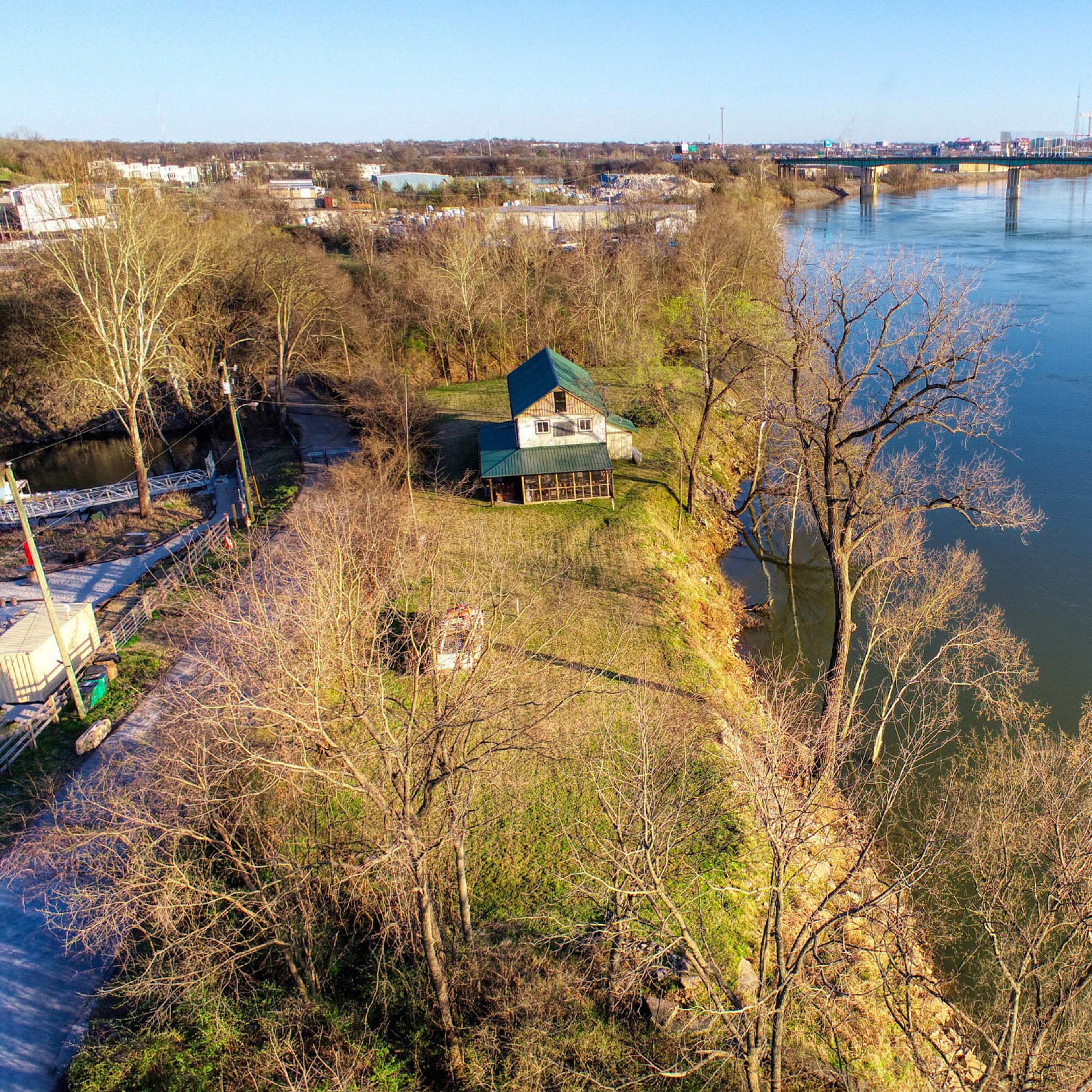 A river flows past a green-roofed house on a wooded bank, with a city skyline visible in the background.