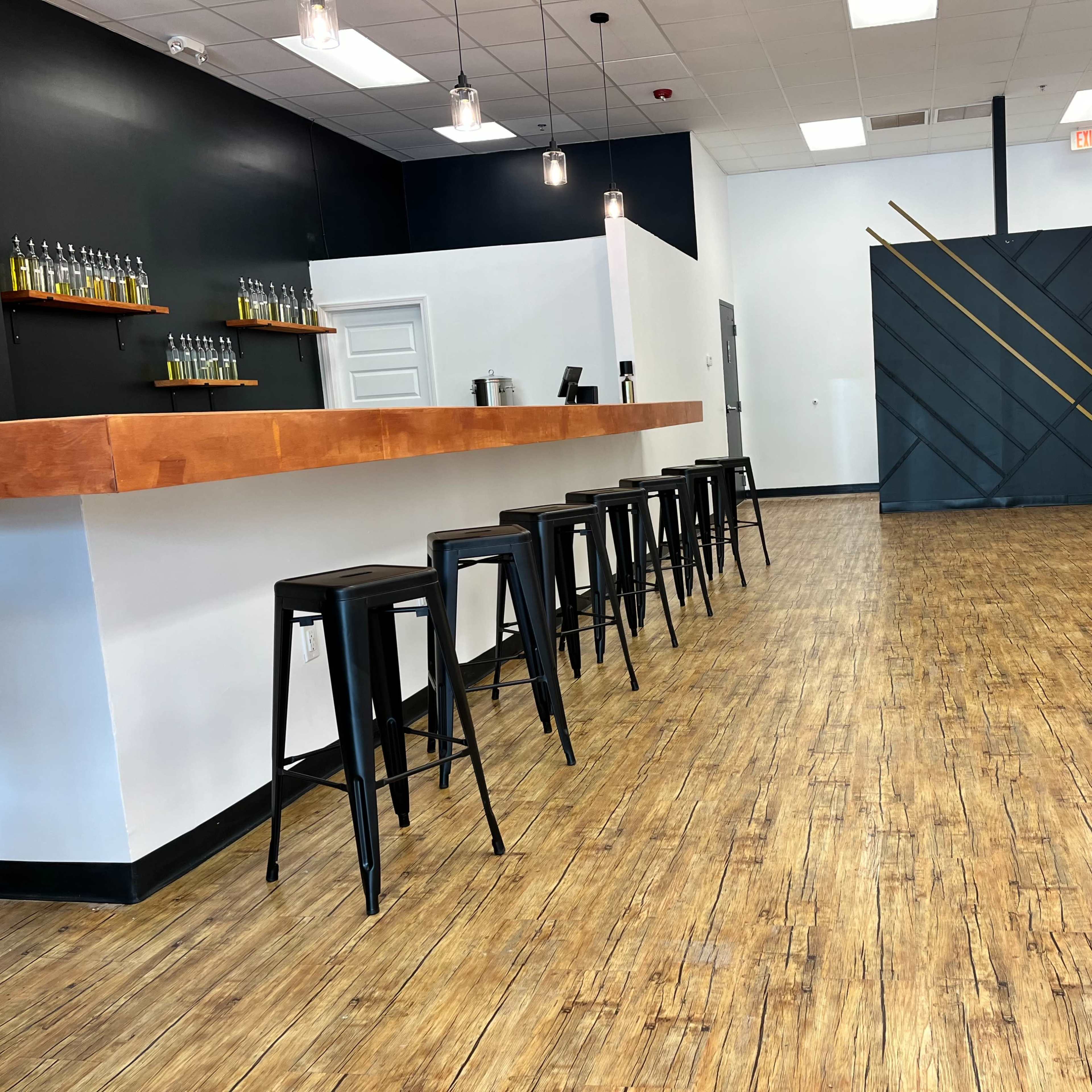 A modern, sparsely furnished interior space with a wooden bar, black metal stools, and a polished wooden floor.