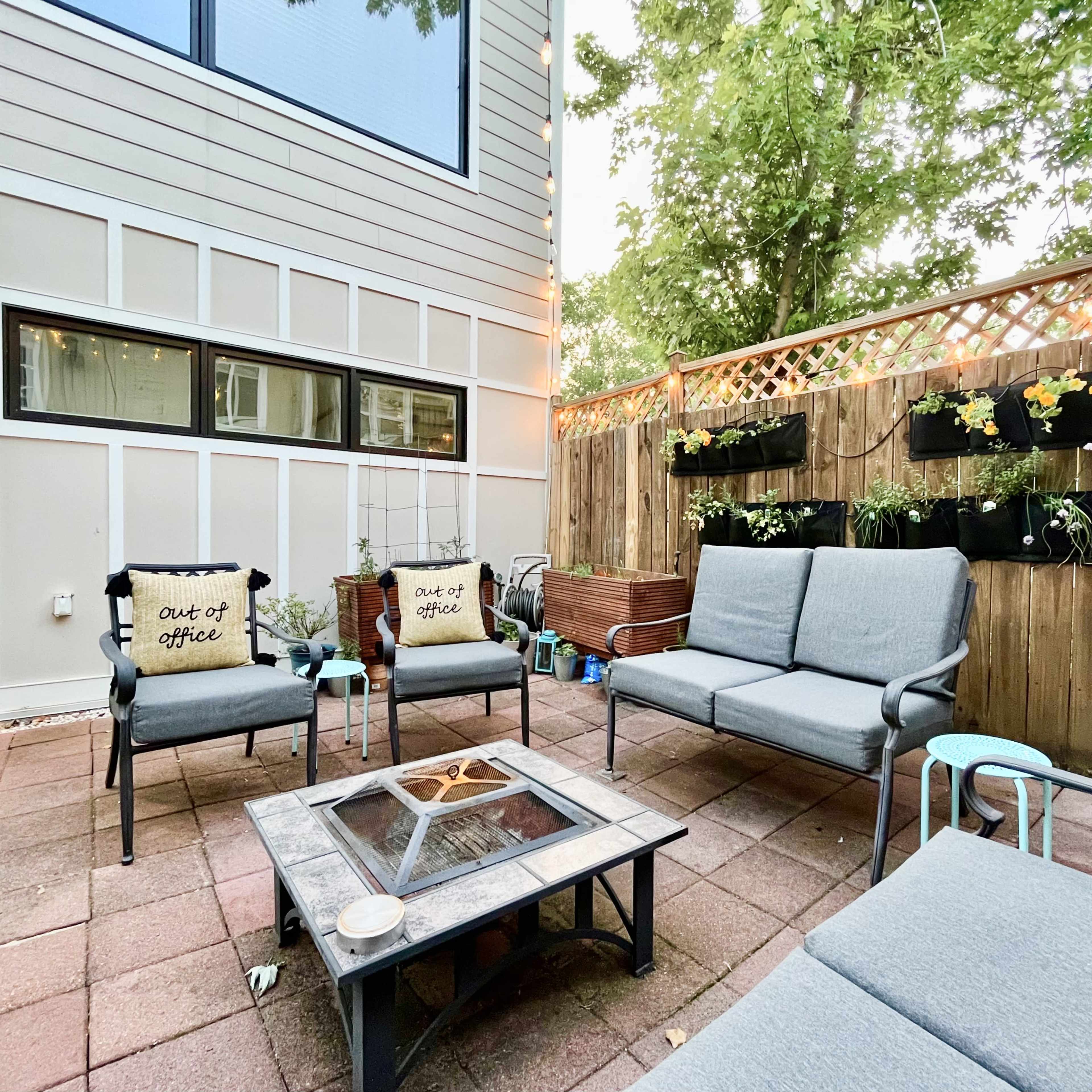 A cozy outdoor seating area features gray couches and a coffee table with a fire pit, surrounded by potted plants and wall-mounted planters.