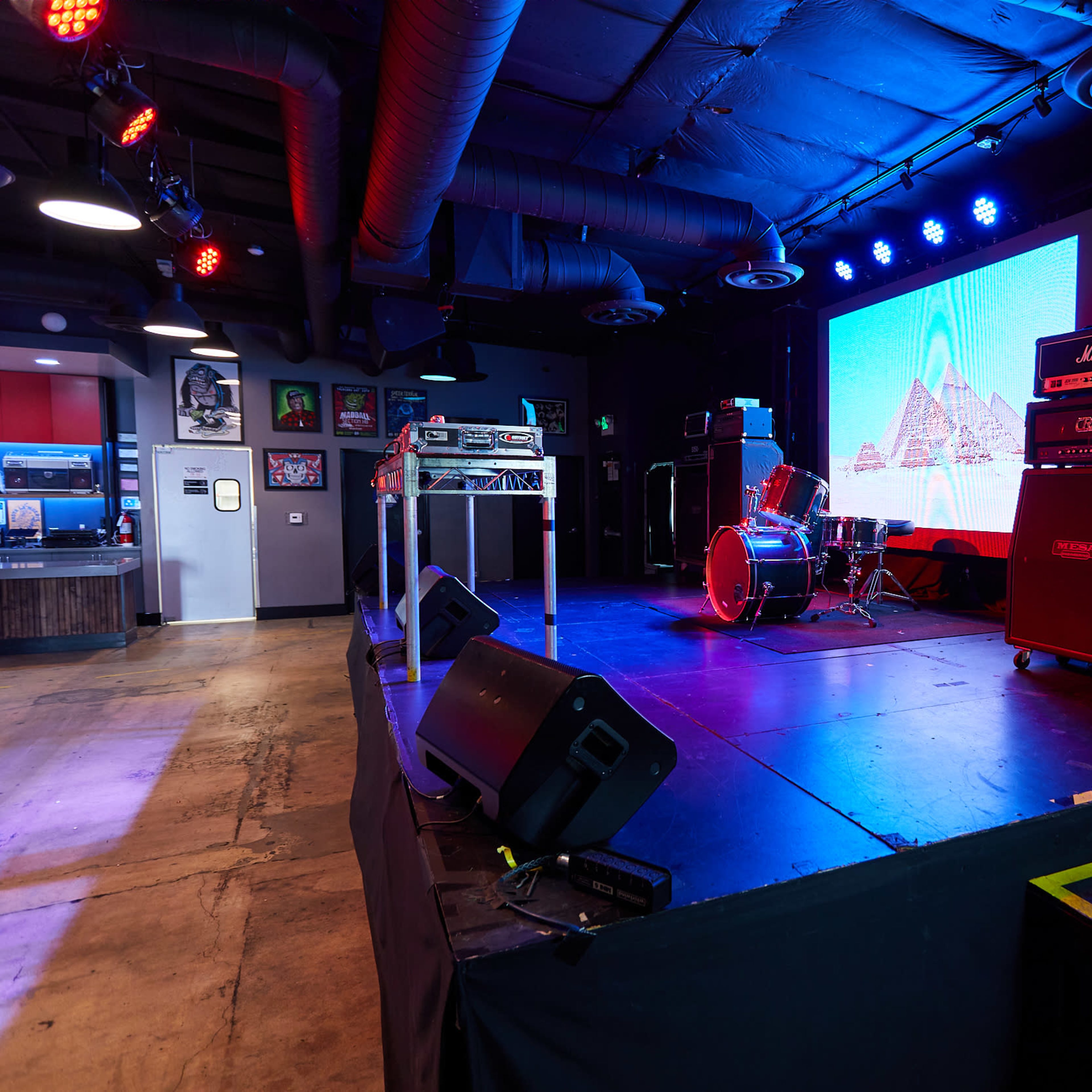 A stage setup with musical equipment, including amplifiers and a drum set, alongside a large screen displaying a visual backdrop.