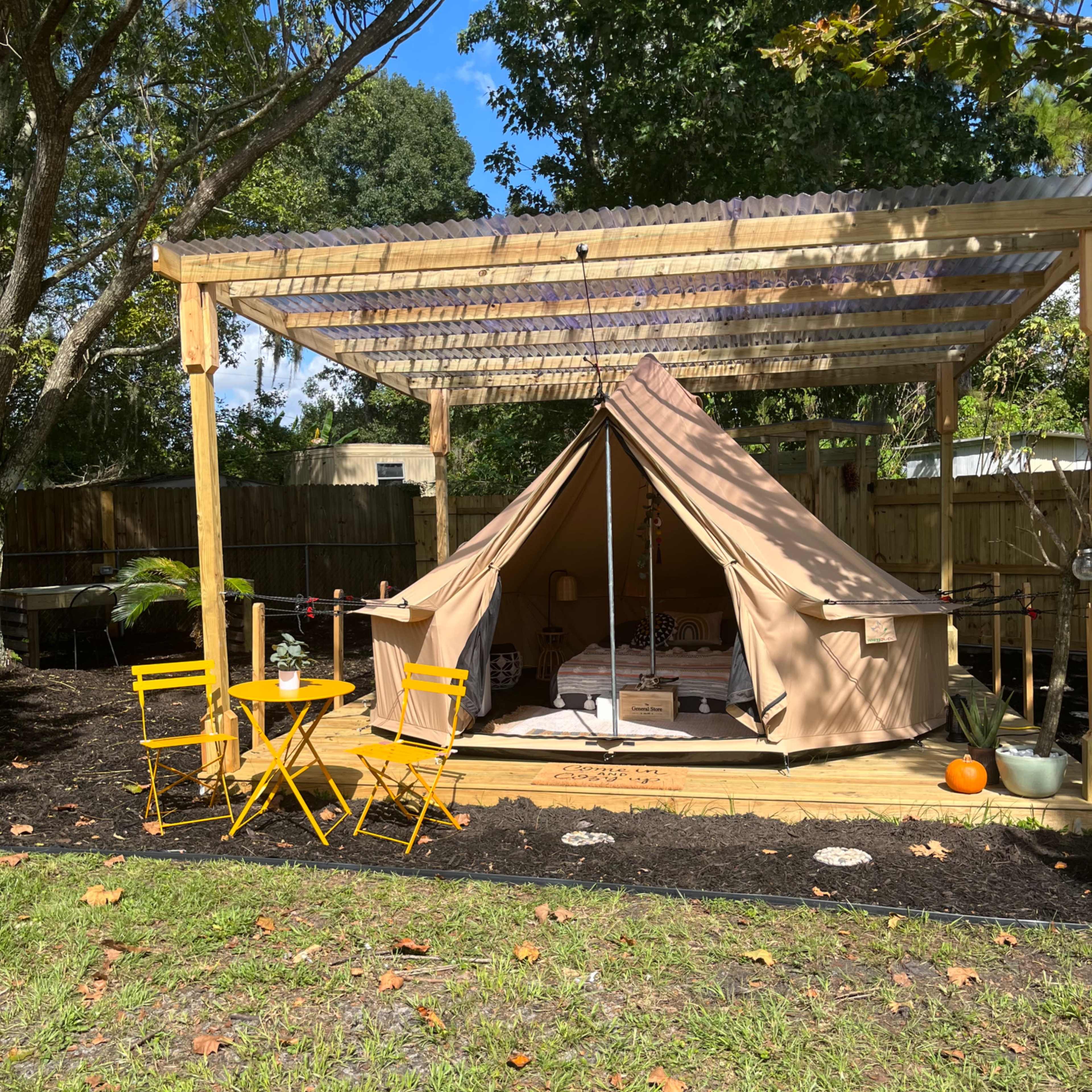 A tent is set up under a wooden pergola, accompanied by yellow chairs on a grassy area, with trees and a fenced background.
