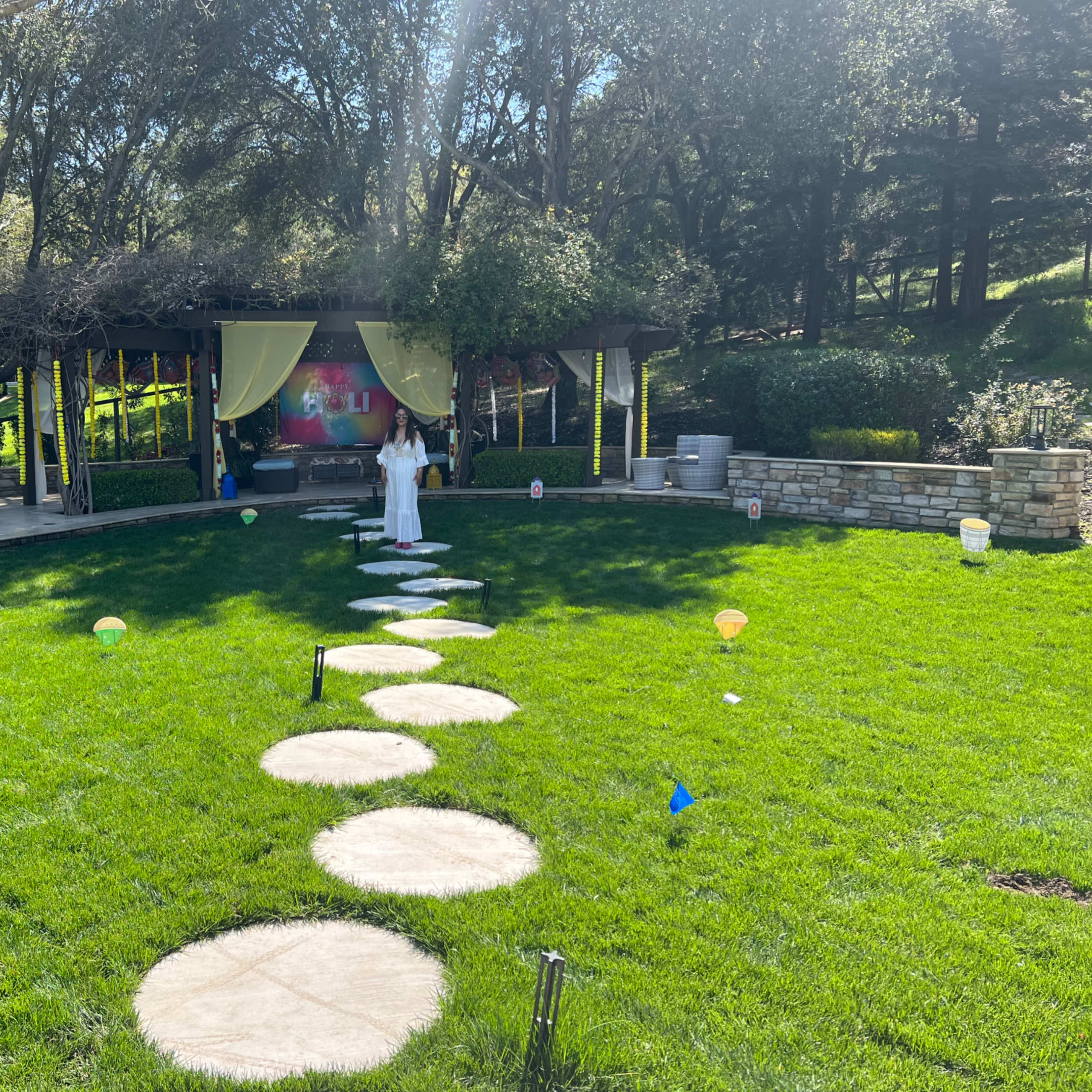 A woman in white stands on a stone pathway leading to a gazebo in a grassy area surrounded by trees.