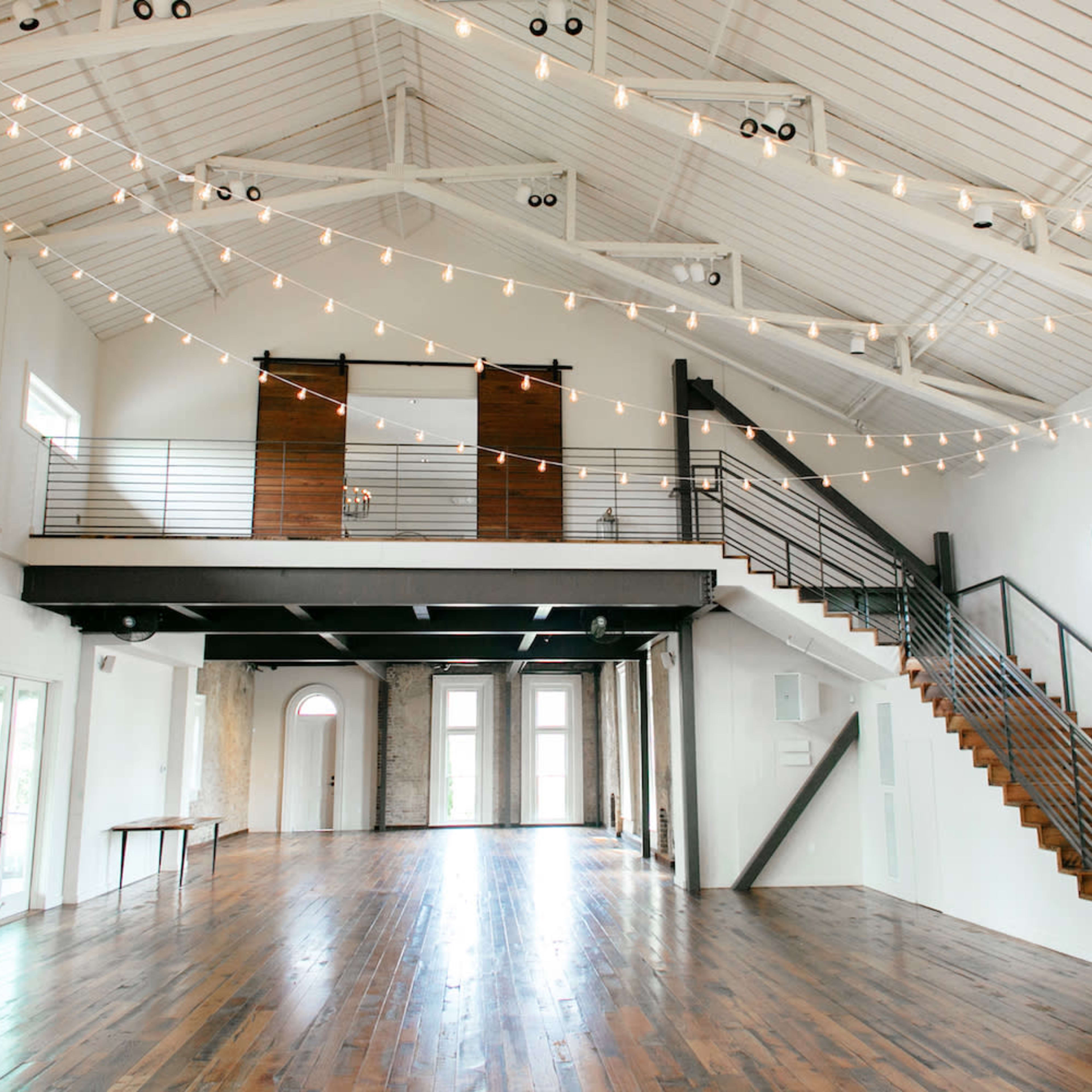 A spacious indoor venue with high ceilings, wooden floors, and string lights, featuring a staircase and large windows.