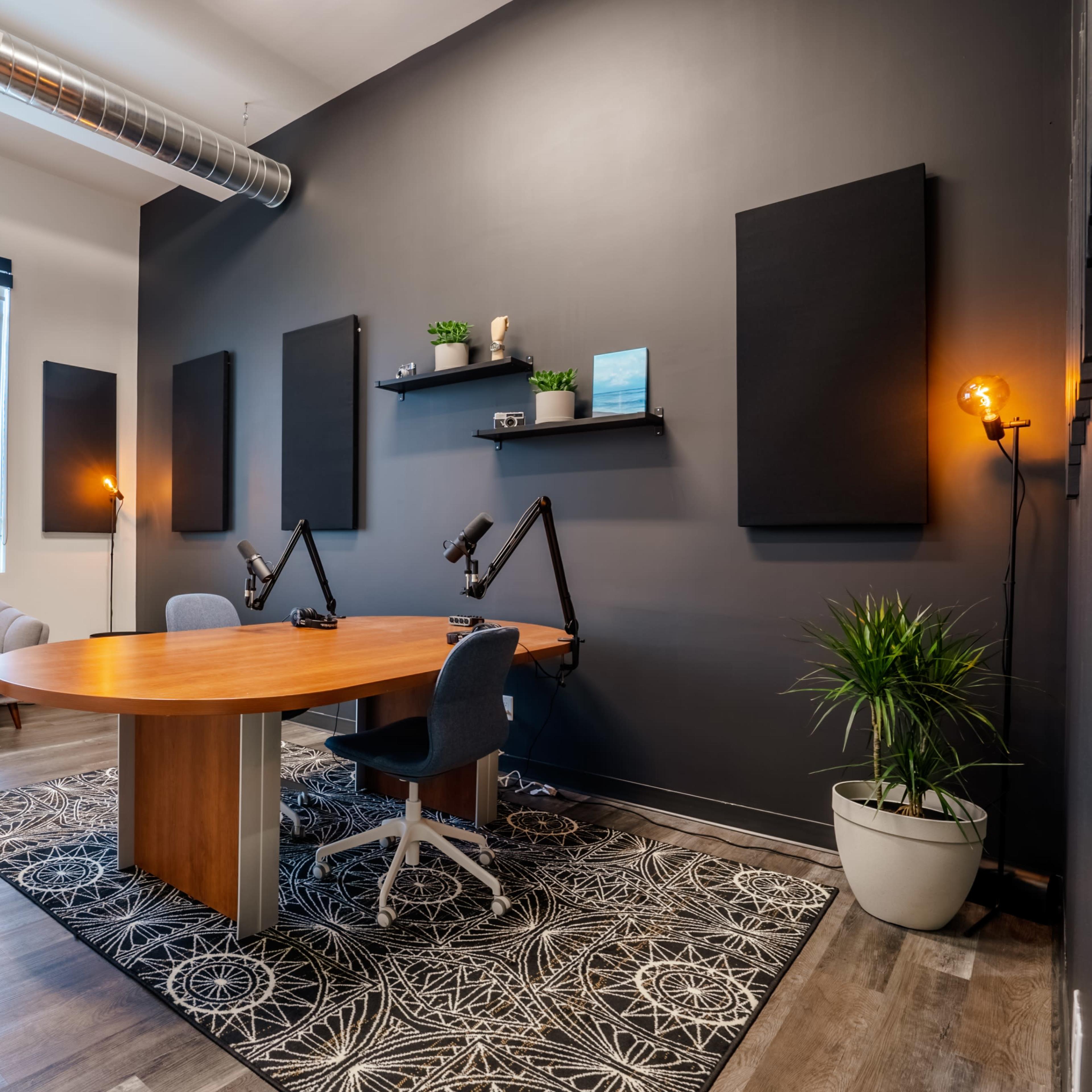 A modern office space features a round wooden table with two adjustable desk microphones, a gray sofa, plants, and black acoustic wall panels against a dark wall.