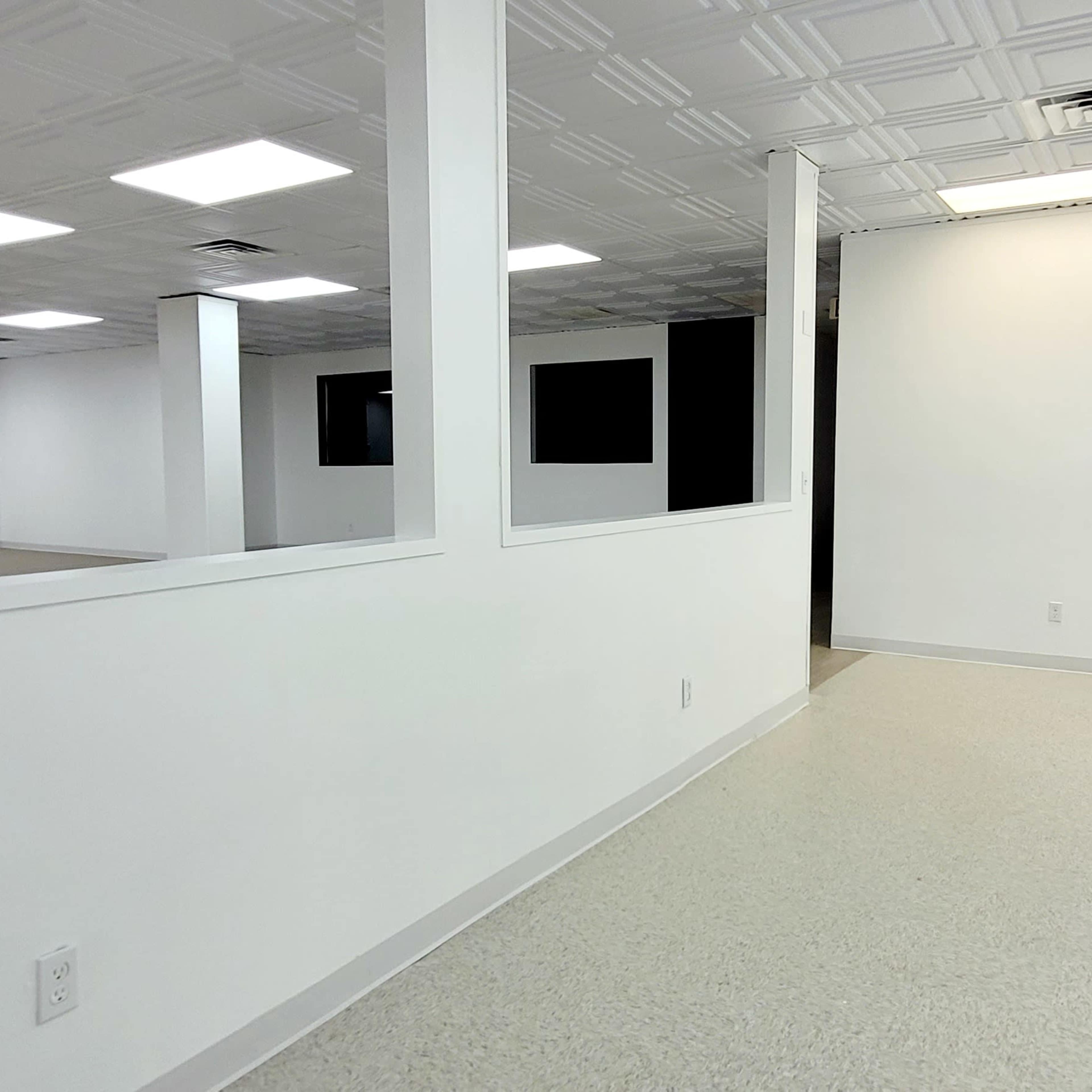 An empty room with white walls, a ceiling with tile patterns, and a light-colored floor, featuring large fluorescent lights and a window that opens to another space.