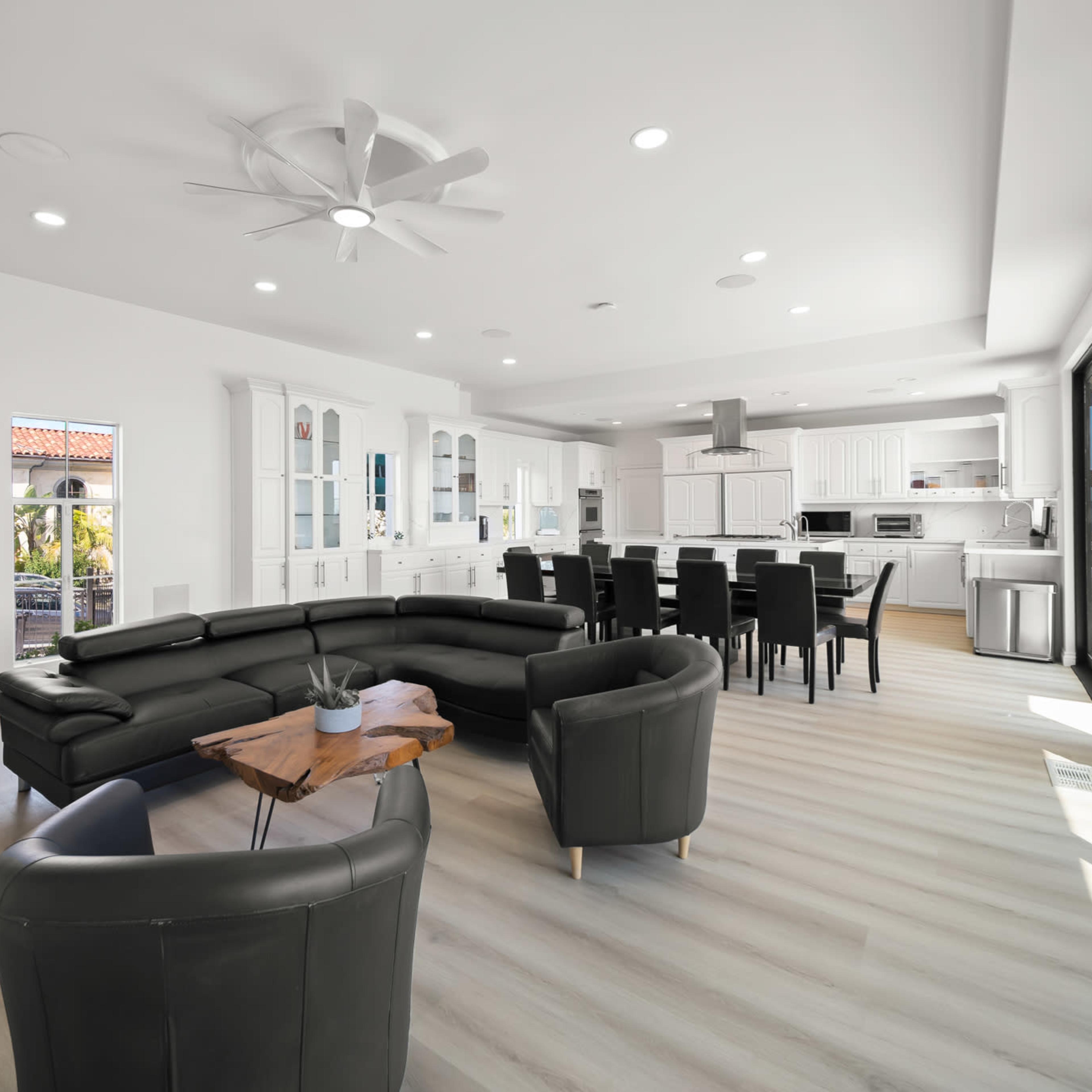 A modern open-concept living space features a black sectional sofa, a wooden coffee table, and a dining area with black chairs adjacent to a bright kitchen.