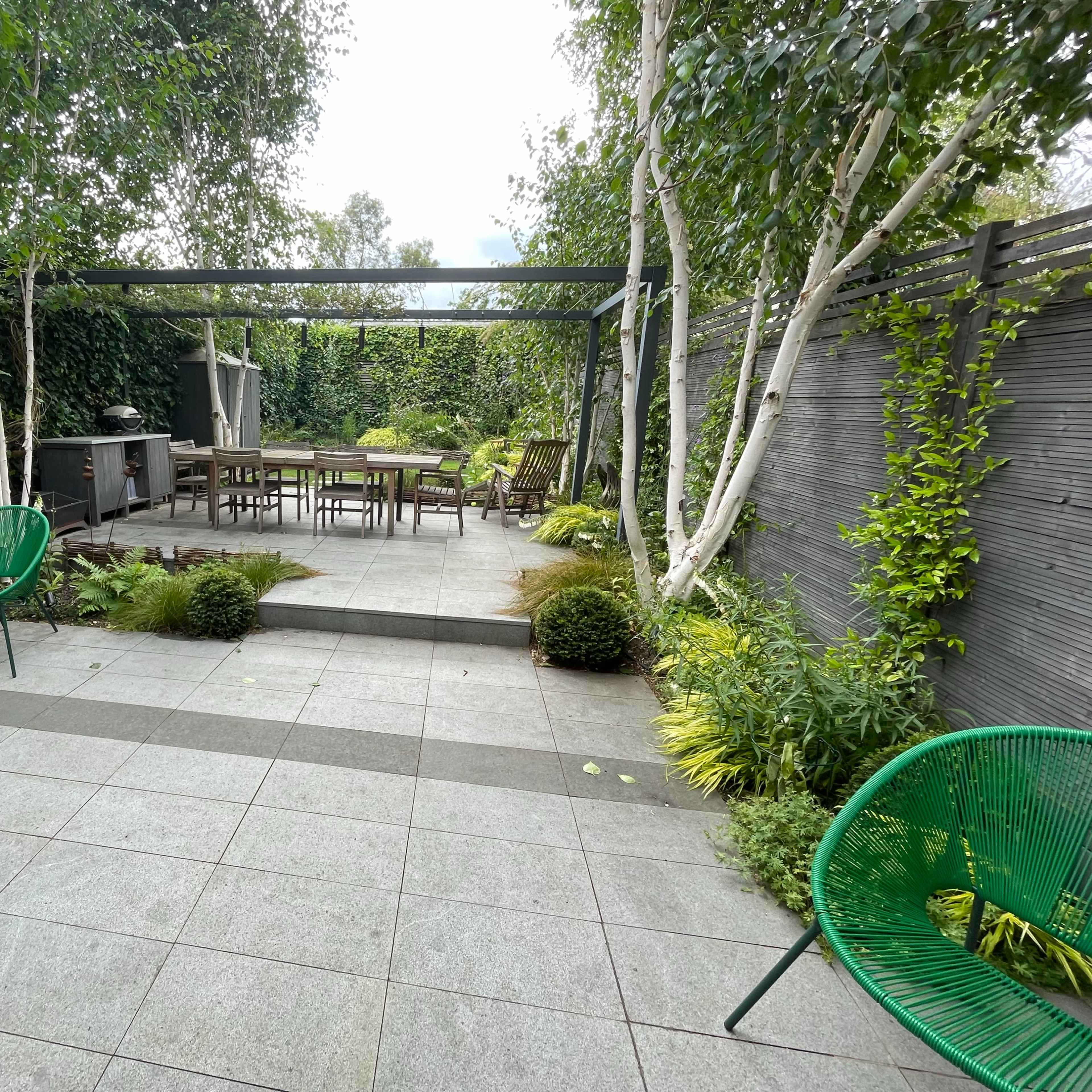 A modern garden features green chairs and a dining table surrounded by lush plants and trees.