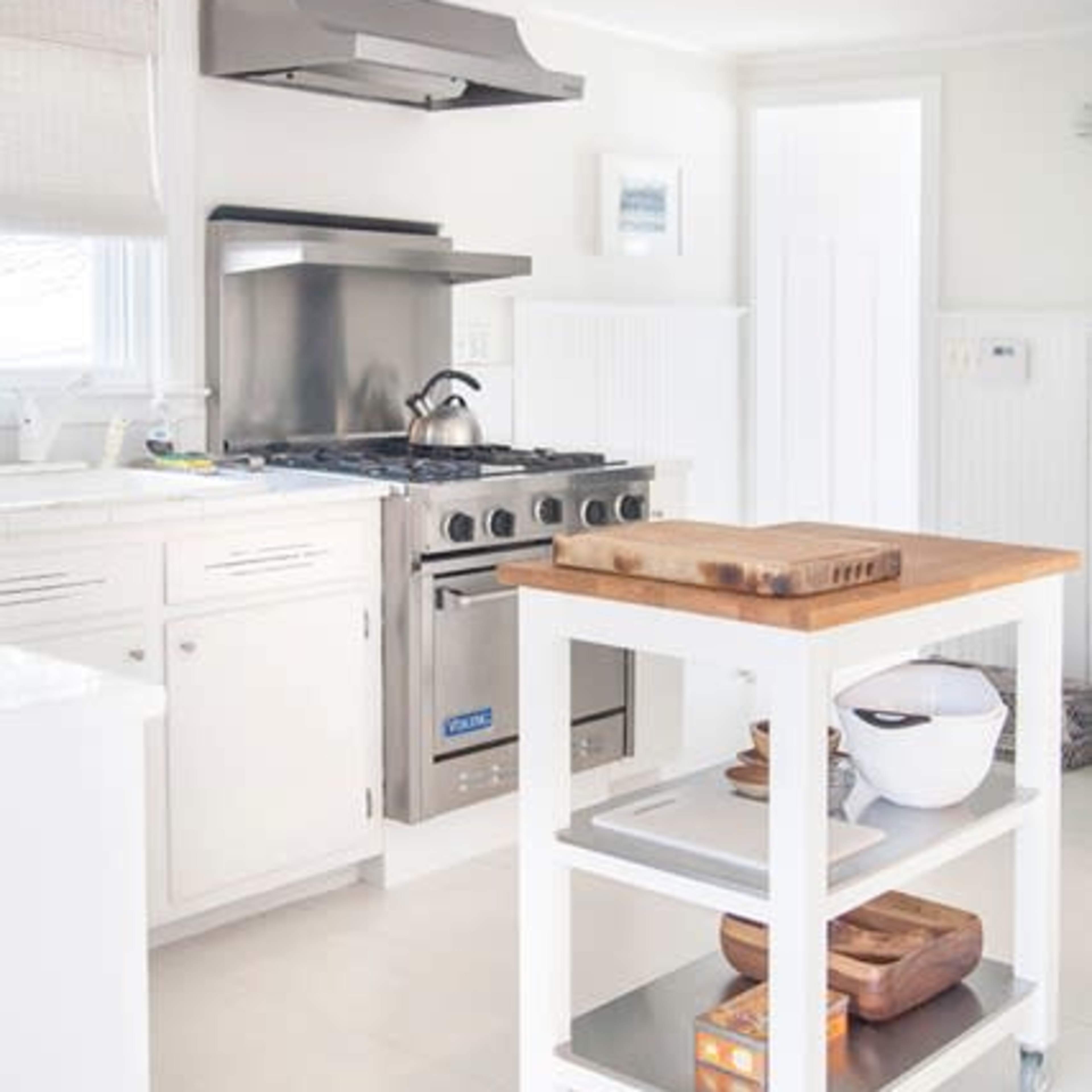 A modern kitchen features white cabinets, a stainless steel range with an exhaust hood, and a central wooden island with open shelving.