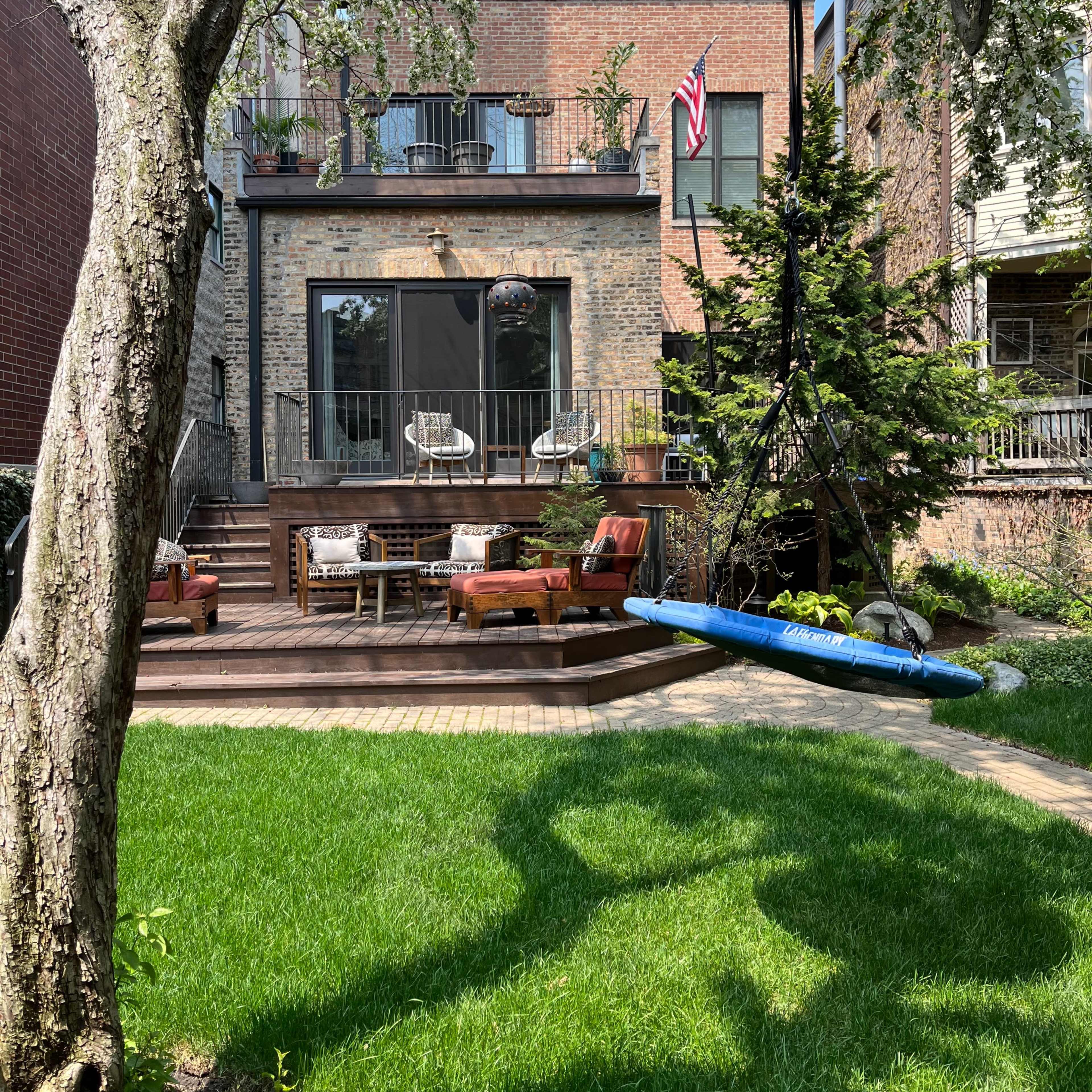 A backyard features a wooden deck with seating and a blue swing hanging from a tree, with a brick building and an American flag in the background.